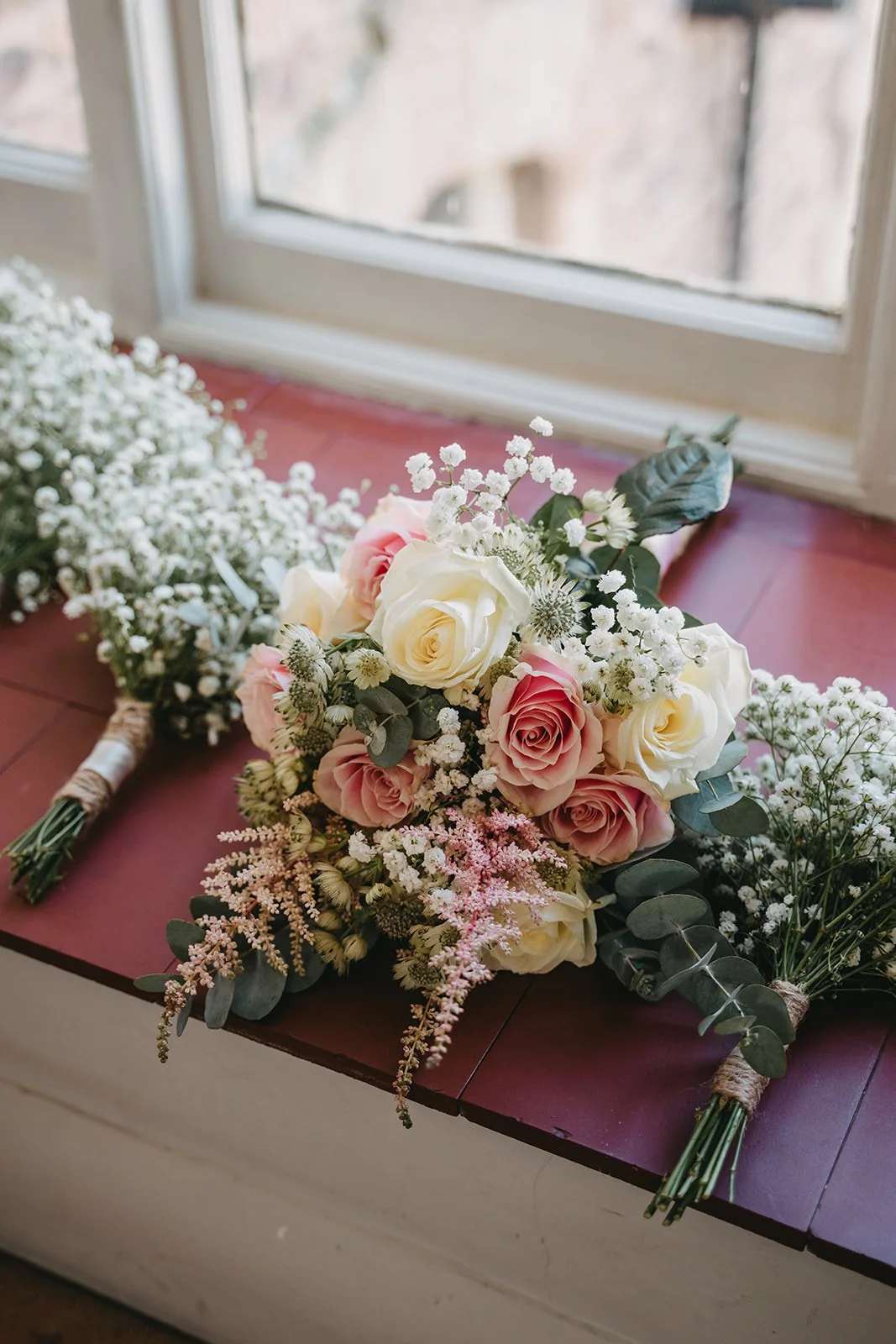 A bouquet of pink and white roses with white baby's breath and greenery resting on a wooden surface near a window.