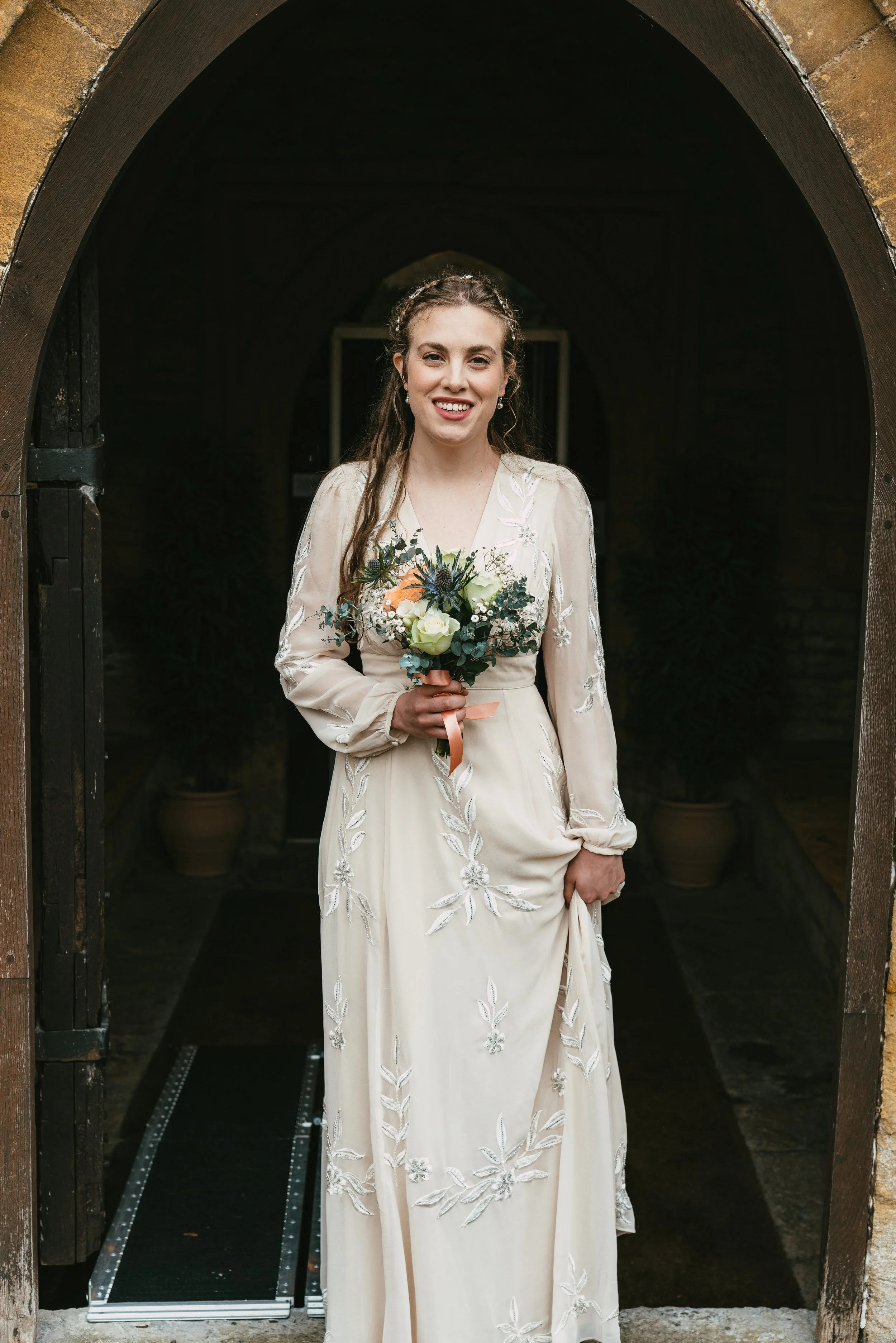 A woman in a cream-colored embroidered dress holding a bouquet of flowers standing in a stone archway.