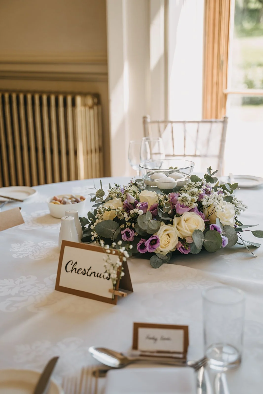 A dinner table decorated with a floral centerpiece of white roses and purple flowers, with a sign labeled 'Chestnut's', set with glassware, plates, and silverware, near a window with natural light.