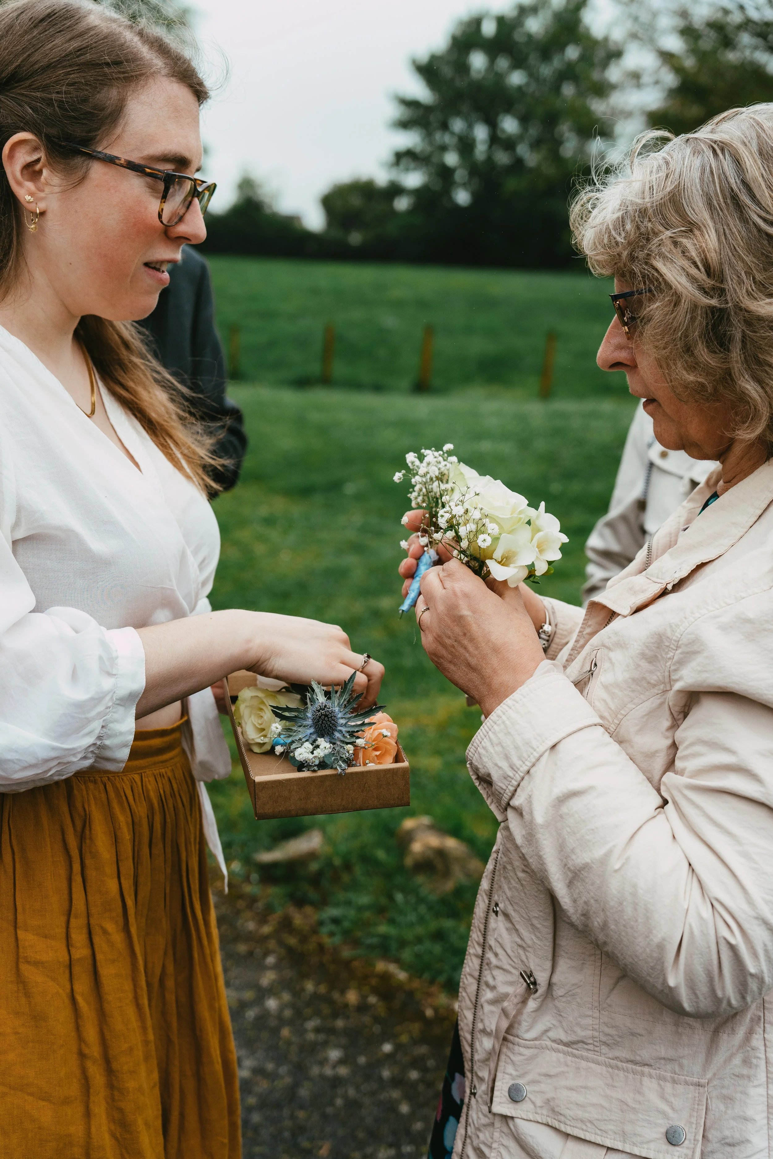 A woman in a white blouse and mustard skirt receives a boutonniere from an elderly woman in a beige jacket at an outdoor event.