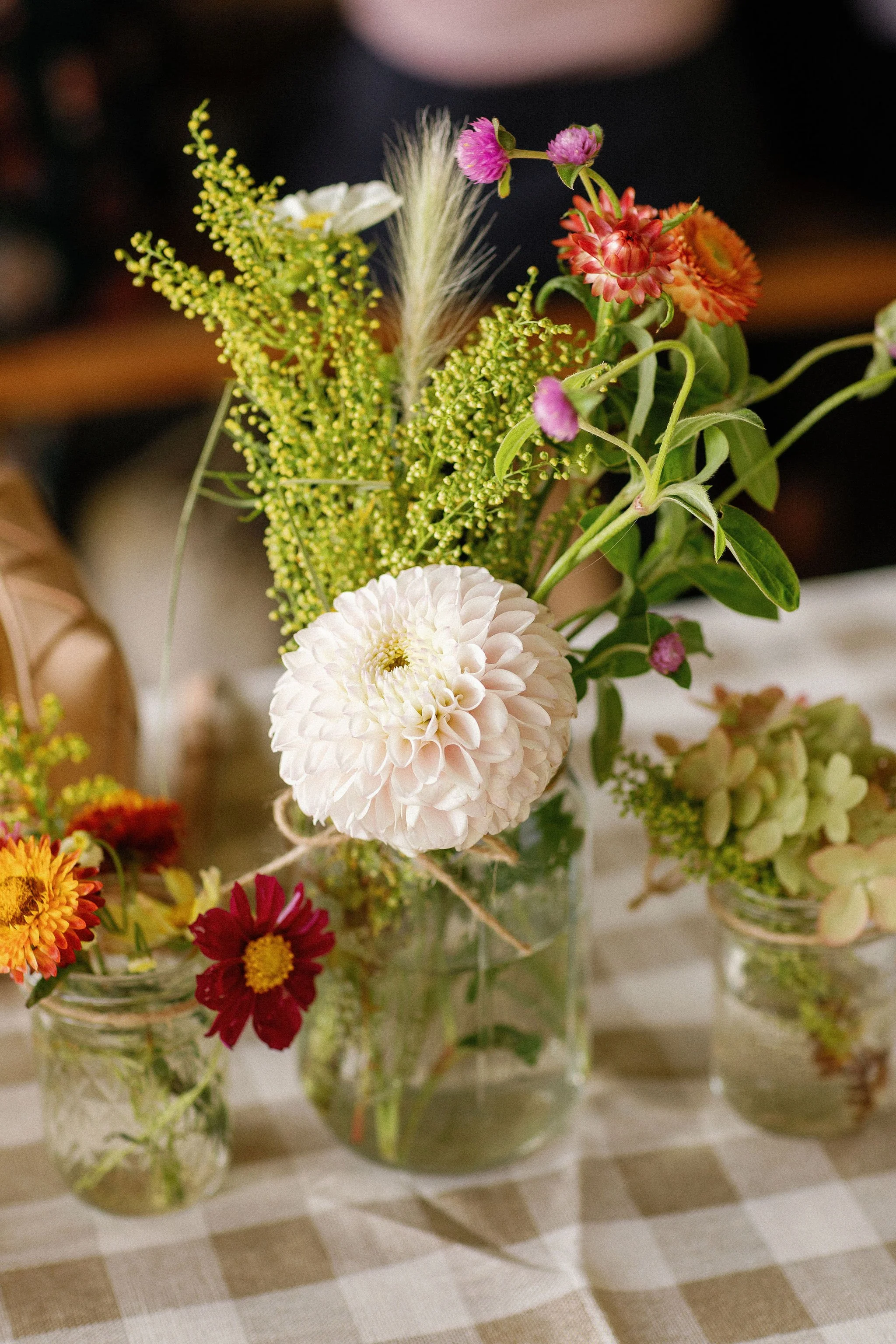 A bouquet of assorted flowers in a glass jar, placed on a checkered tablecloth, with smaller flower arrangements nearby.