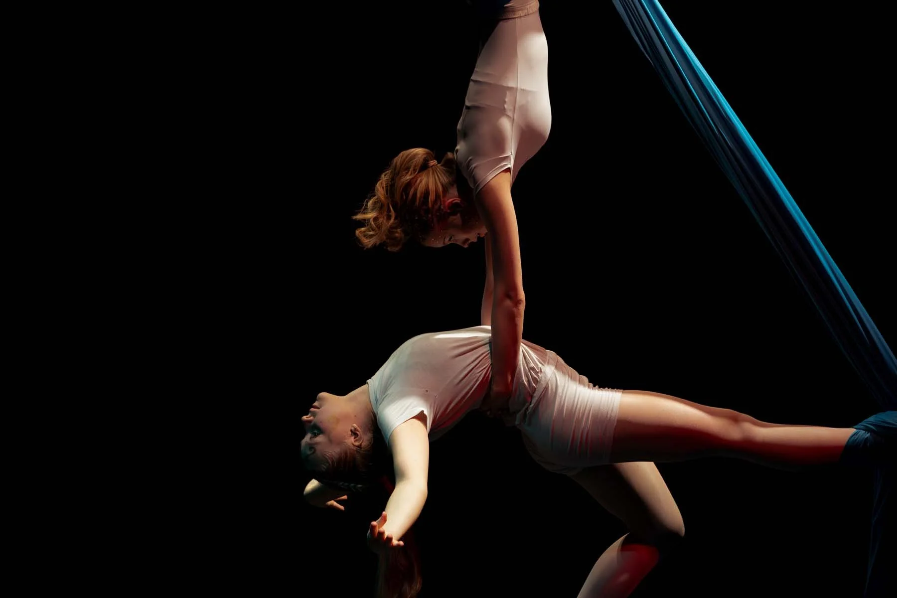 Kaksi naista tekee akrobaattista esitystä, toinen tukeutuu toisen selkään ja toinen on kallistuneena ylöspäin, taustalla musta.