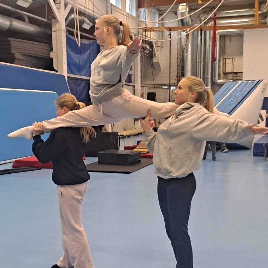 Kolme tyttöä harjoittelee akrobatiaa salissa, toinen seisoo ja pitää kiinni toisen jalkoja, joka istuu ilmassa,"