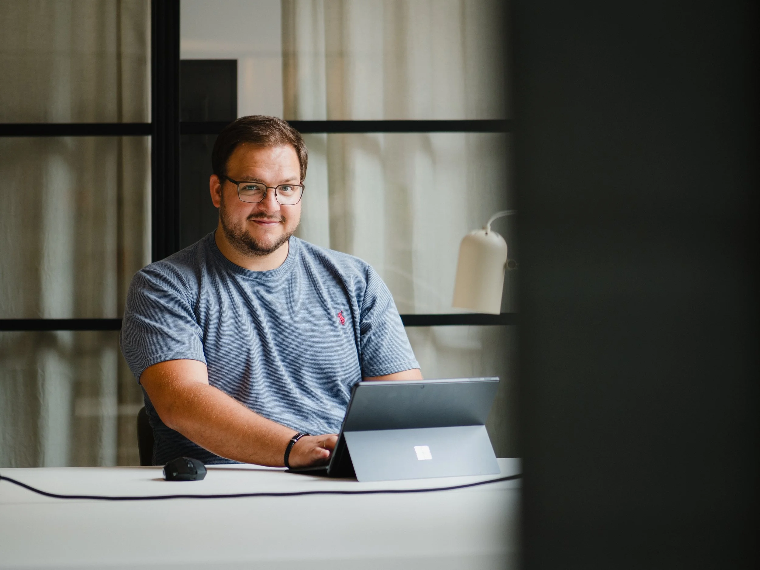 Ein Mann mit Brille sitzt an einem weißen Tisch vor einem Laptop, im Hintergrund sind Fenster und eine weiße Lampe sichtbar.