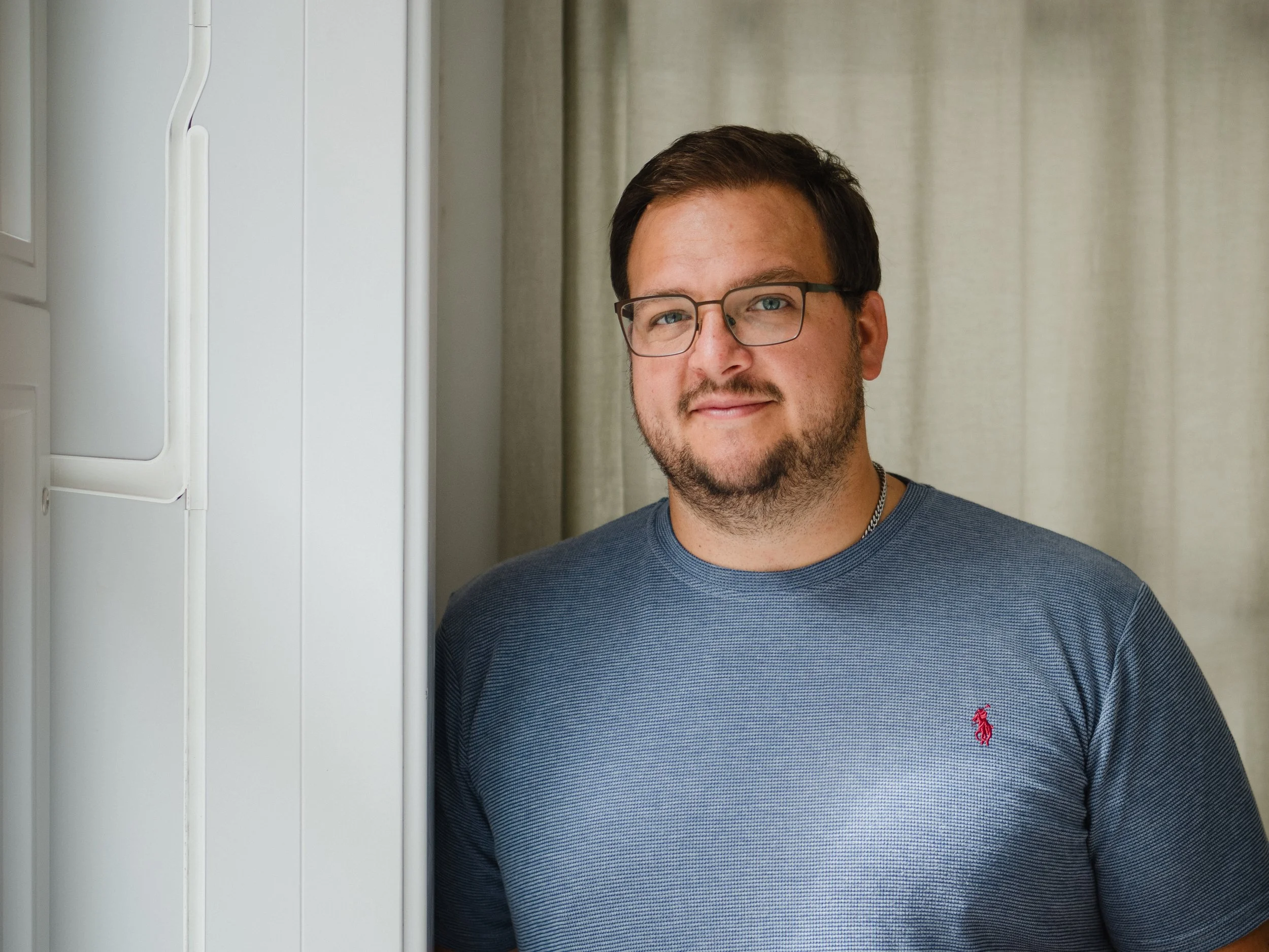 Ein Mann mit Brille und Bart trägt ein graues T-Shirt und steht vor einem hellen Vorhang in einem Raum.