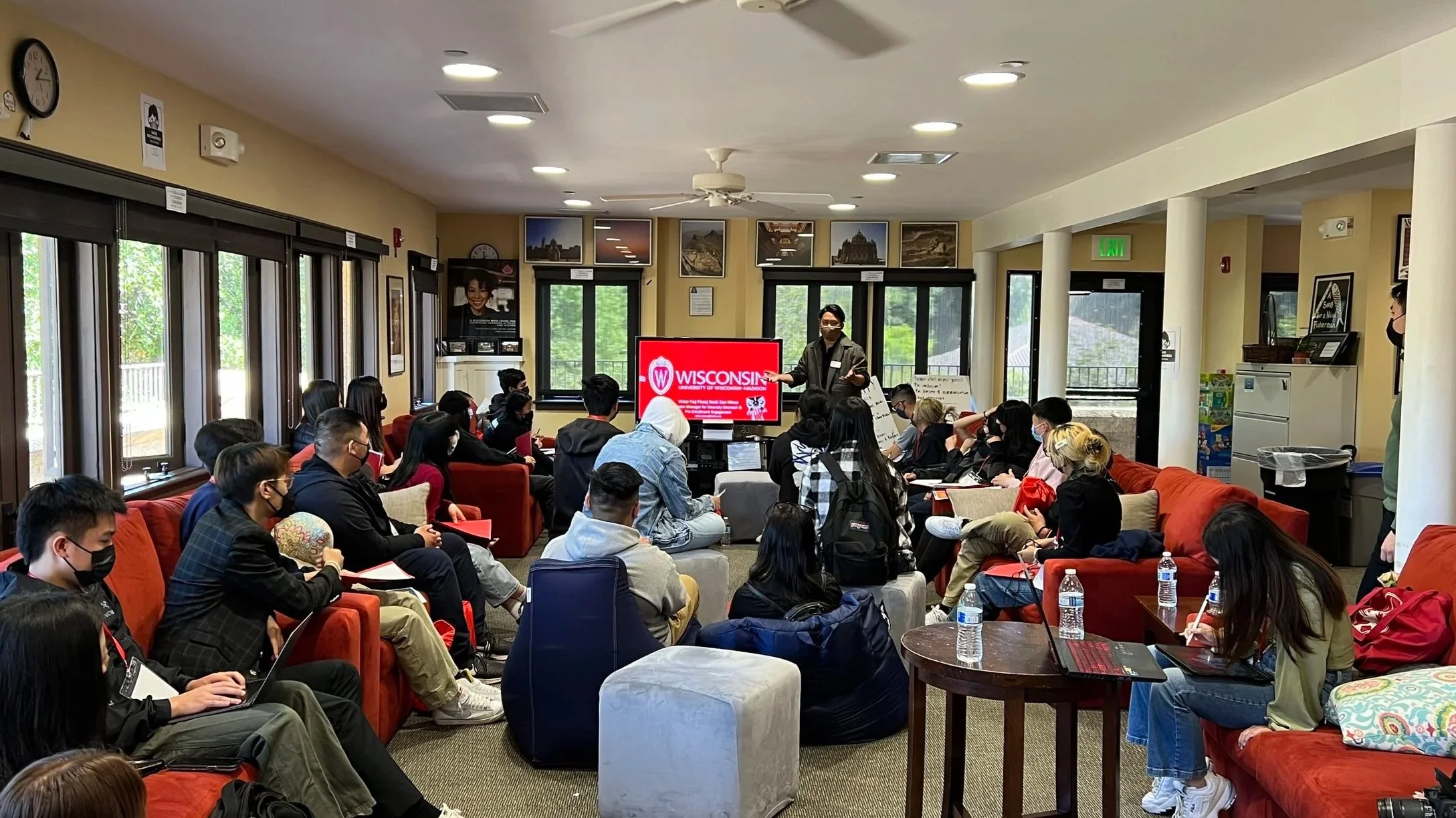 A group of students attending a presentation in a bright room with large windows, sitting on red and gray sofas and chairs, listening to AASPIRES' Executive Director, Vince, standing in front of a large screen.