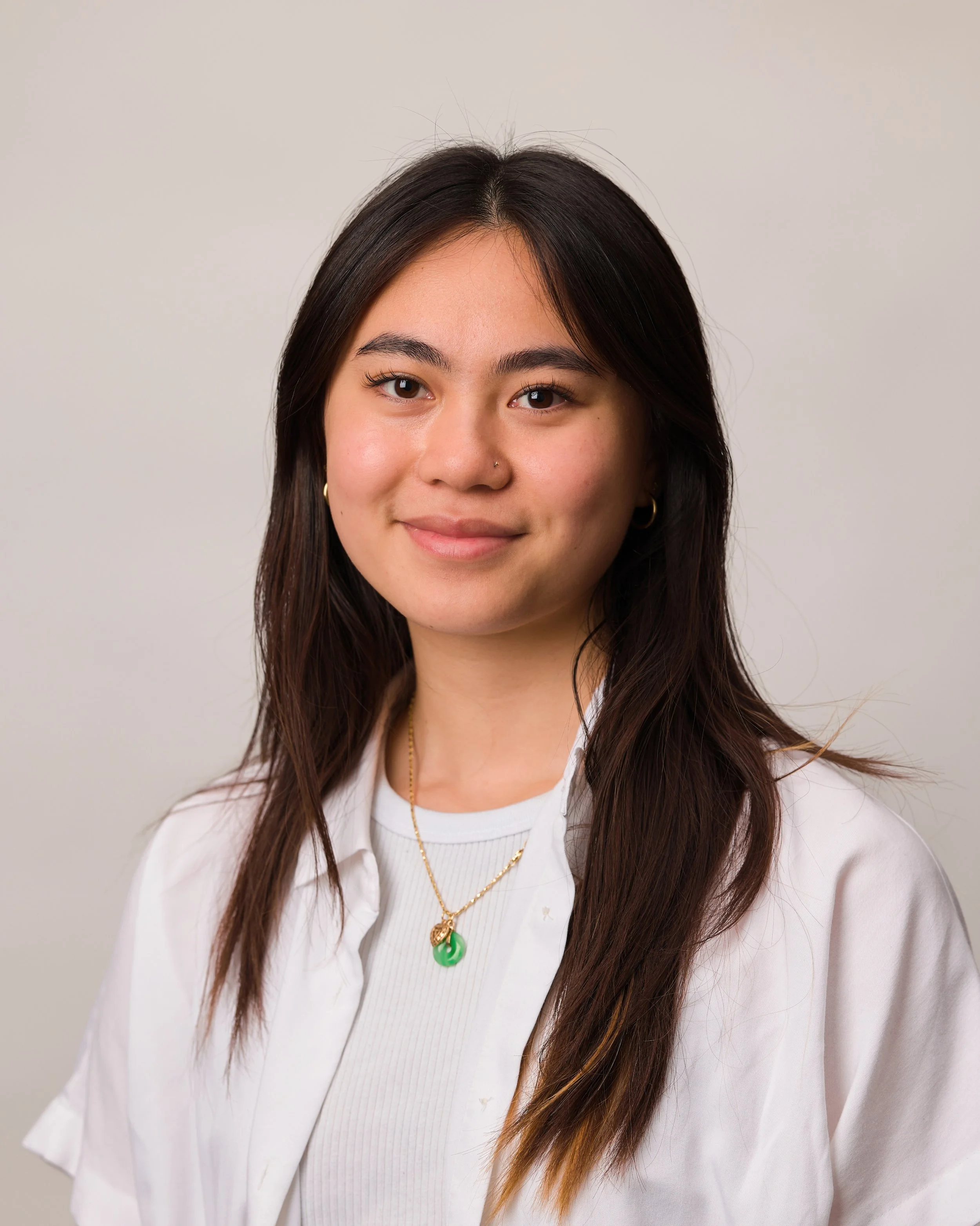 A young woman with long dark hair wearing a white shirt, gold jewelry, and a green pendant necklace, smiling at the camera against a plain light-colored background.
