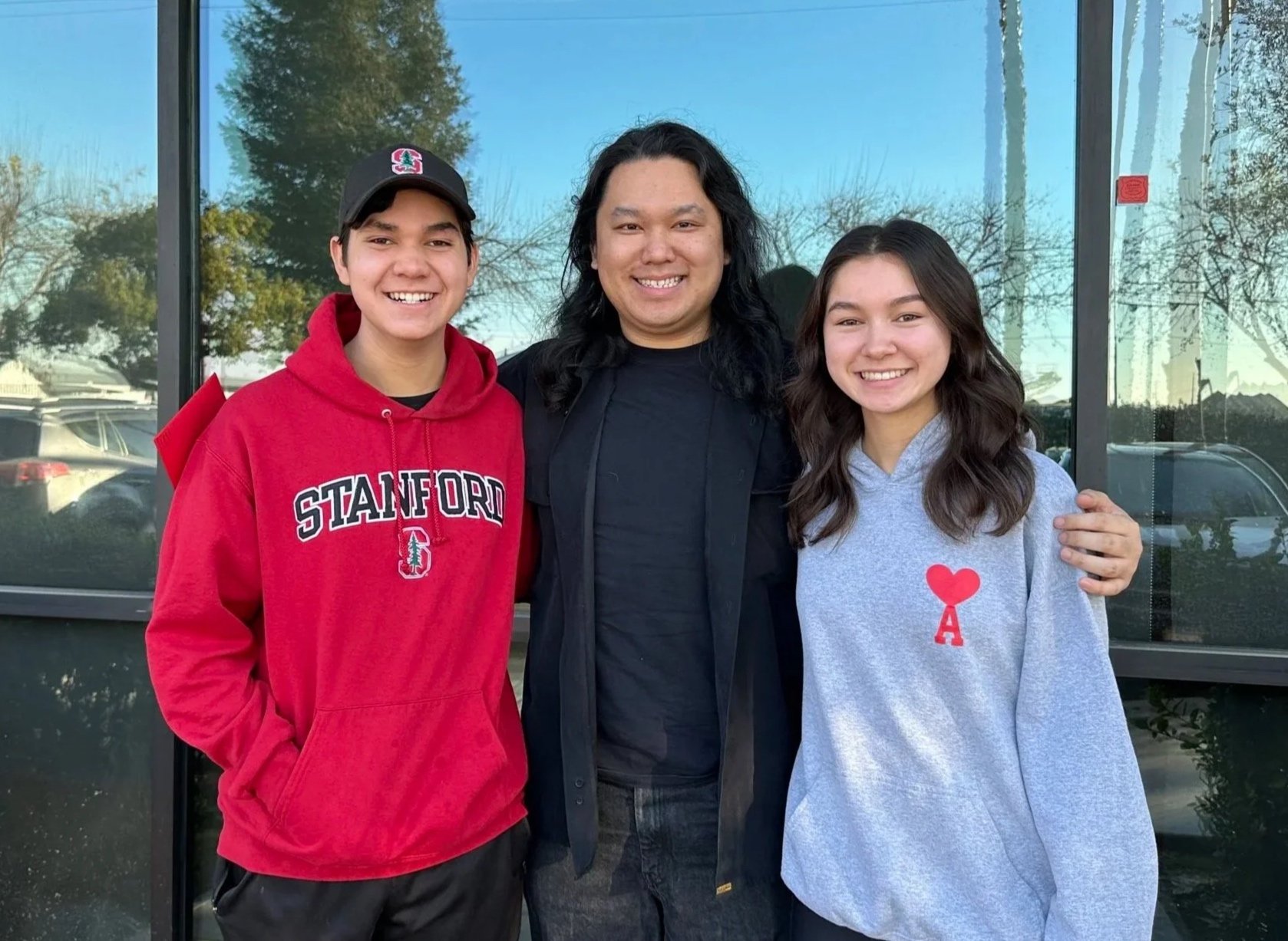 Three smiling people standing outside in front of a glass window. The person on the left is wearing a red Stanford hoodie and a black cap. The person in the middle has long black hair and is wearing a black shirt and jacket. The person on the right is wearing a gray hoodie with a red heart and letter A. The background shows parked cars and trees.