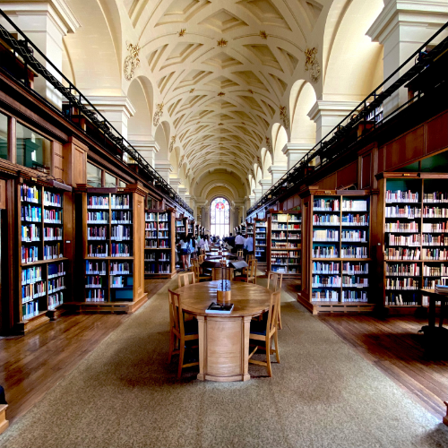 Inside a grand library with high vaulted ceilings, wooden bookshelves filled with books lining both sides, a long wooden table with chairs in the center, and large windows at the far end.