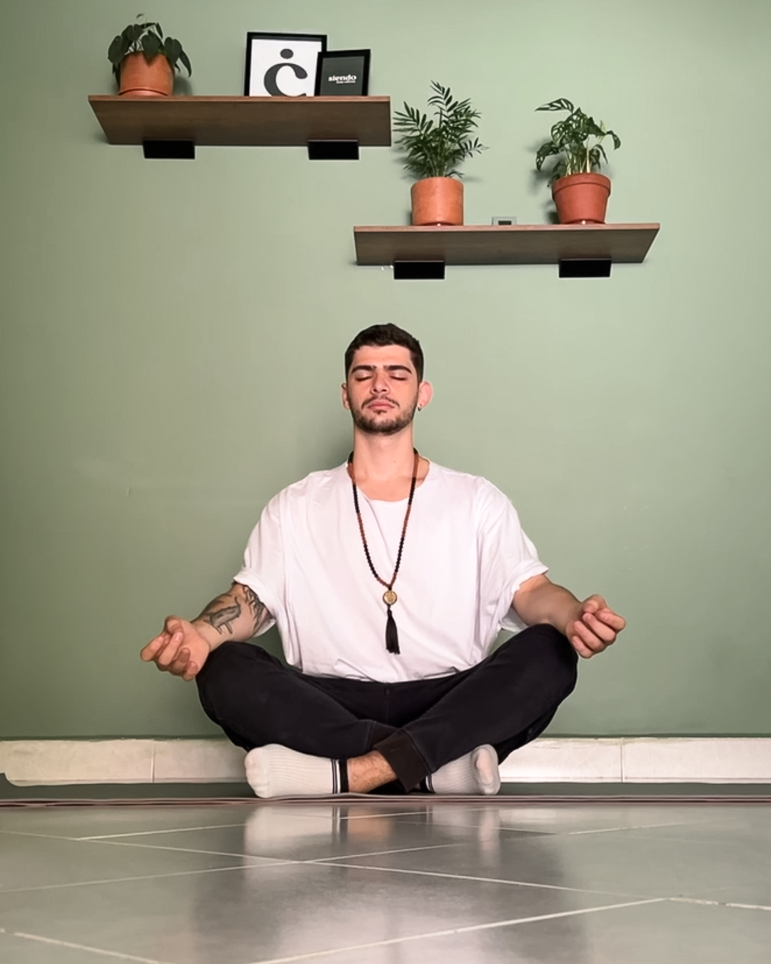 Manuel Marín meditando en posición de loto en un suelo de baldosas gris en una habitación con paredes verdes, con estantes con plantas y cuadros en la pared.