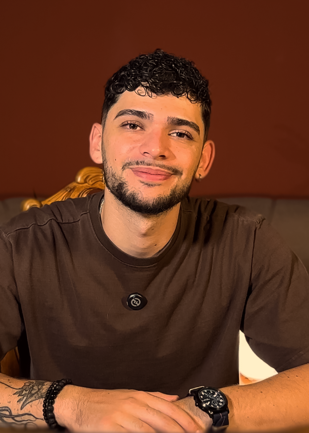 Joven con barba, con cabello rizado, viste una camiseta marrón y un reloj negro, sonriendo frente a un fondo marrón.