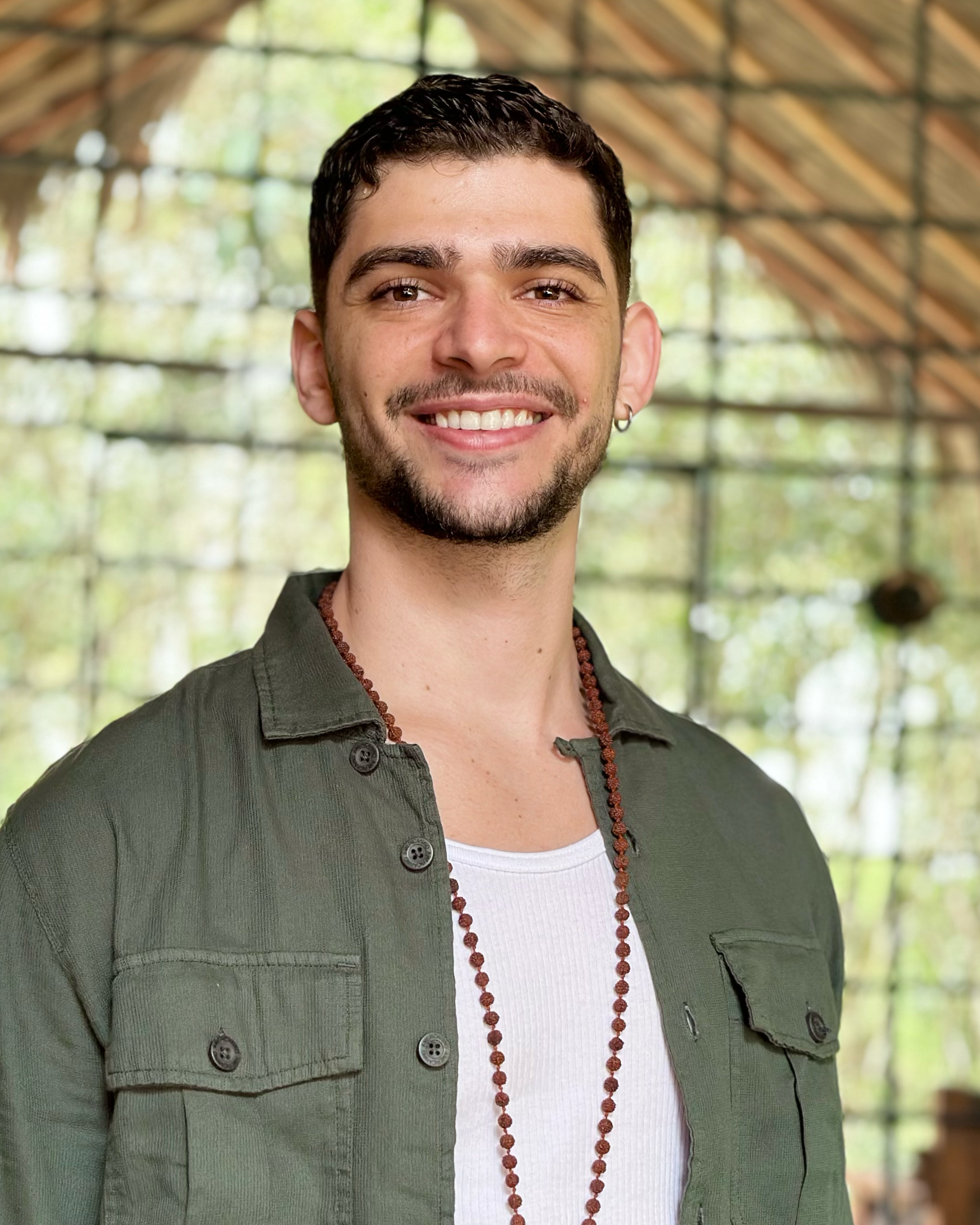 Manuel Marín sonriendo con barba, camiseta blanca y chaqueta verde en un espacio con estructura de madera y ventanas grandes con vista a árboles.