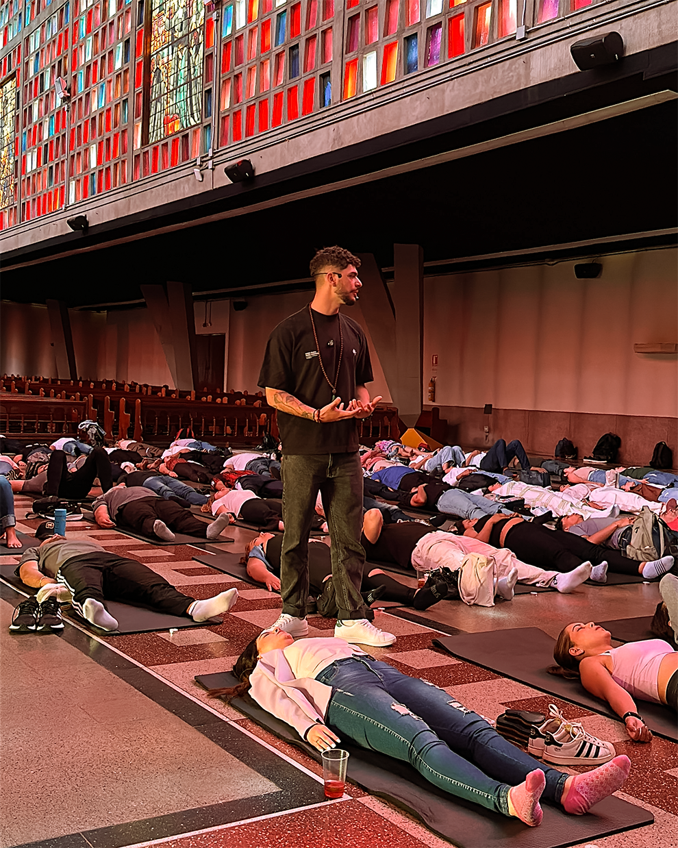Grupo de personas participando en una clase de yoga o meditación en un auditorio con ventanas de colores. Algunas personas están acostadas en colchonetas en el suelo, mientras que una persona de pie (Manuel Marín) los guía en la actividad.