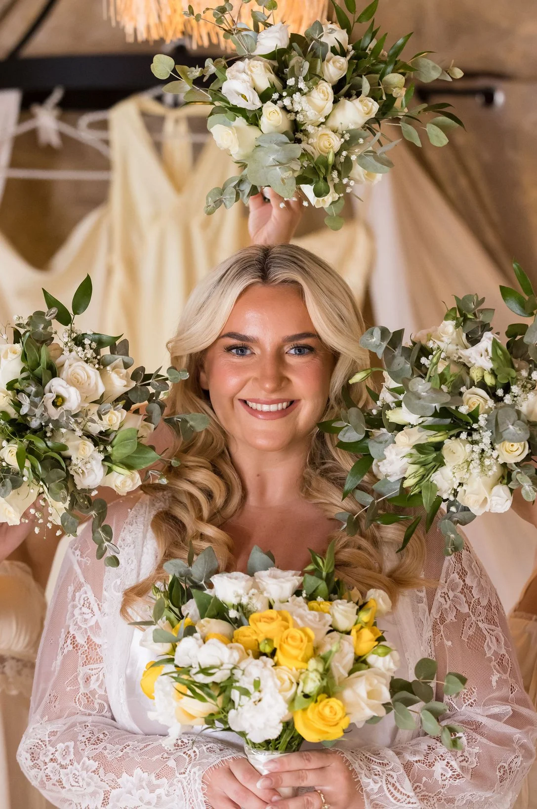 A smiling bride holding a bouquet of yellow and white roses with greenery, surrounded by other women holding similar bouquets, in a room decorated with rustic and floral decor.