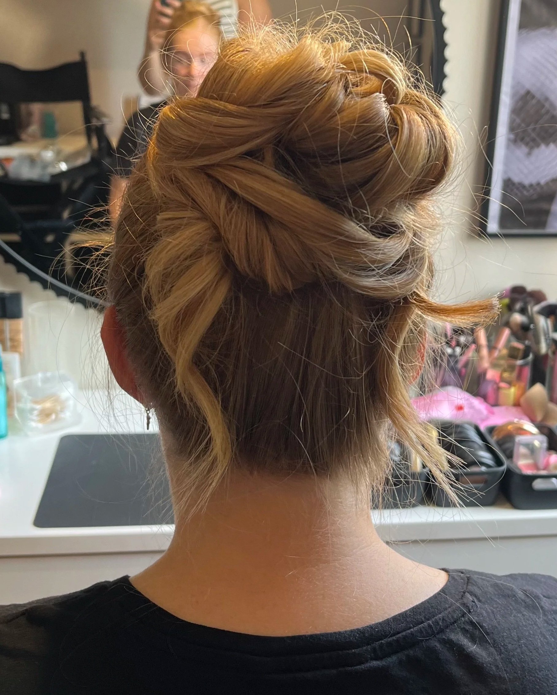Back of a woman with an intricate updo hairstyle, surrounded by hair styling tools and brushes, in a salon setting.