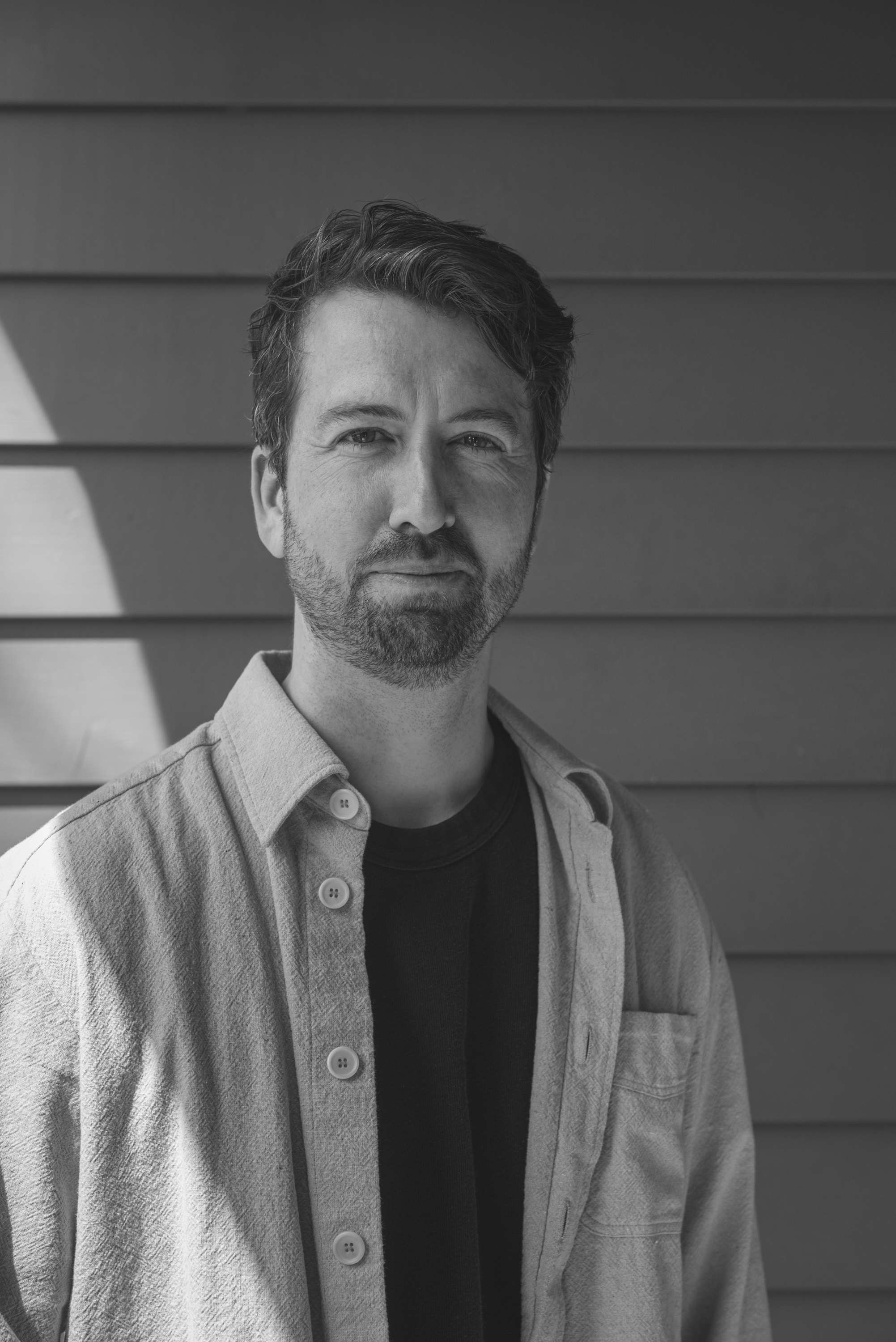 Black and white portrait of a man with wavy hair and facial hair, wearing a button-up shirt over a T-shirt, standing in front of horizontal siding.
