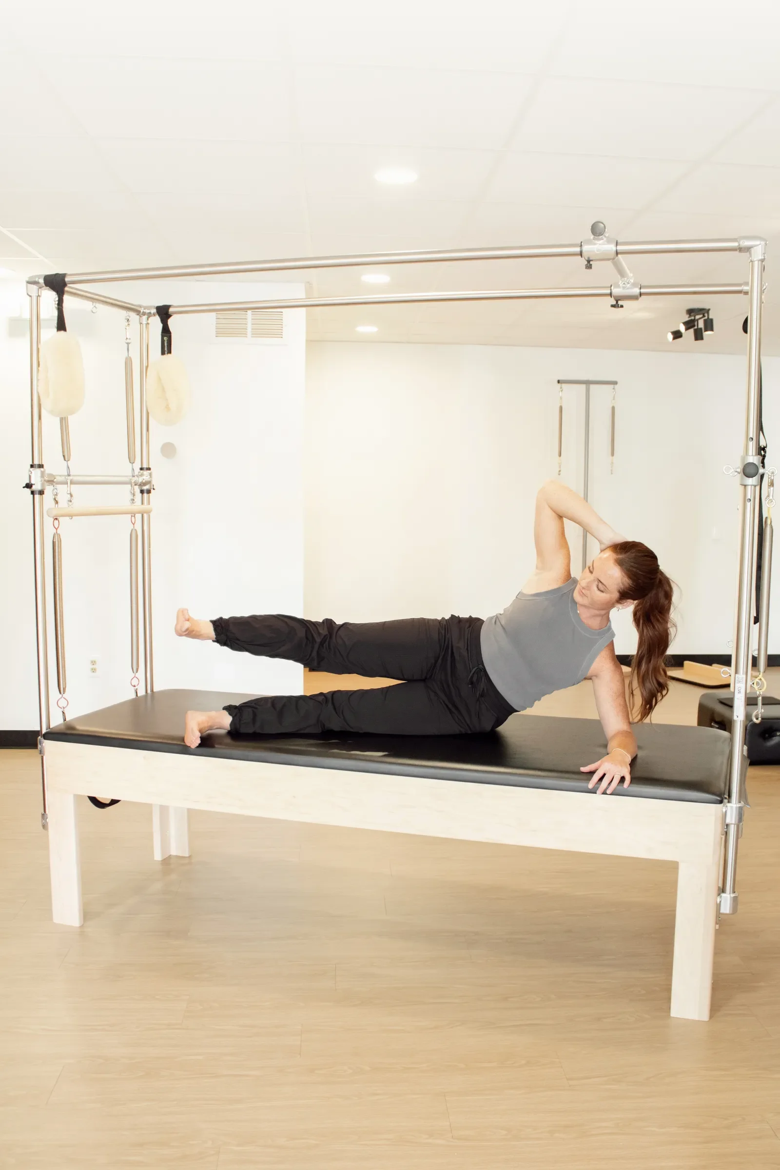 A woman is practicing Pilates on a reformer machine in a studio, lying on her side with one arm supporting her head and the other arm resting on the platform, wearing a gray shirt and black pants.
