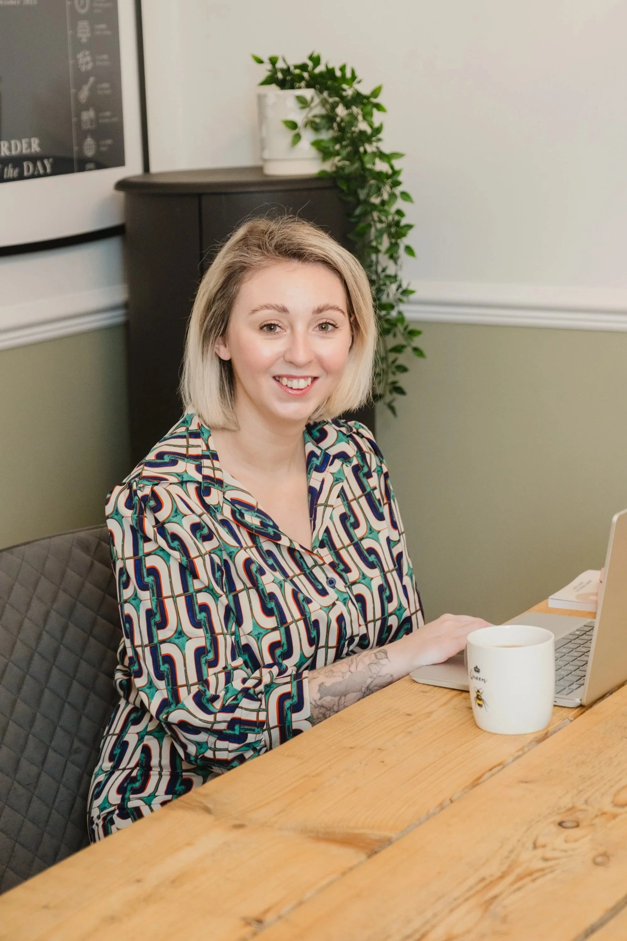 Cassie Marns, Founder of Marns Marketing, sits at a wooden table, smiling at the camera with a laptop in front of her, a white coffee mug nearby, and a green plant behind her in a white pot, in a well-lit indoor setting.