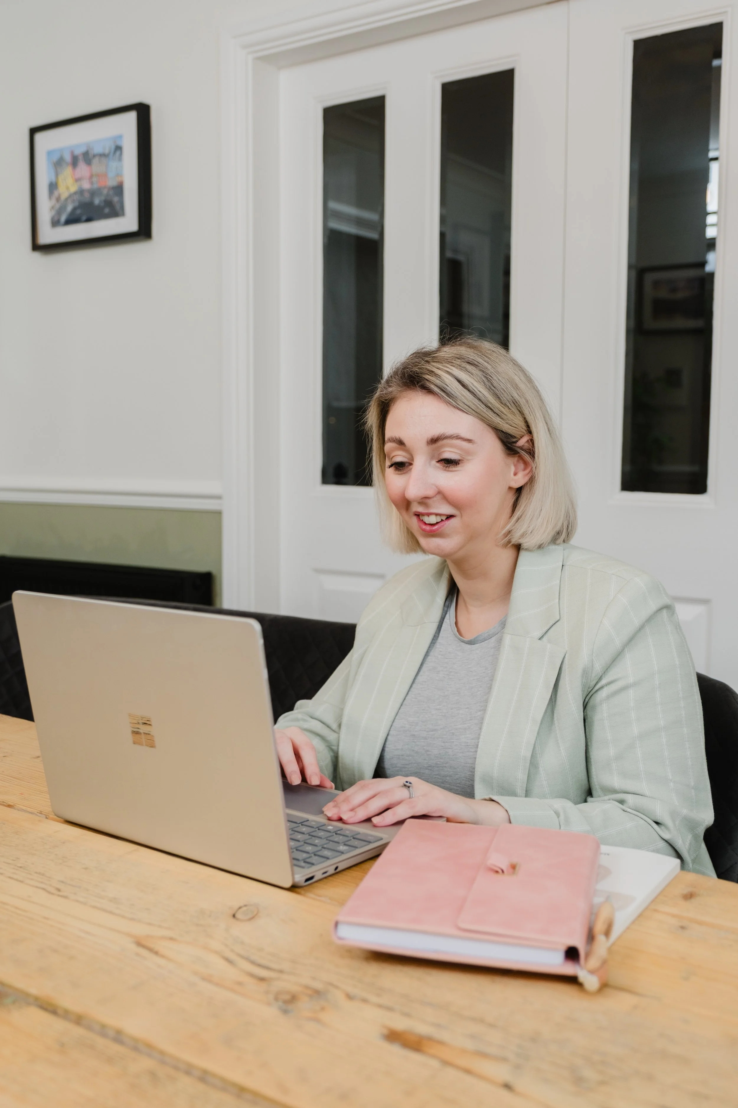 A woman with blonde hair, wearing a light-colored striped blazer and a gray t-shirt, smiles while working on a silver laptop at a wooden table in a room with white walls and glass-paneled double doors.