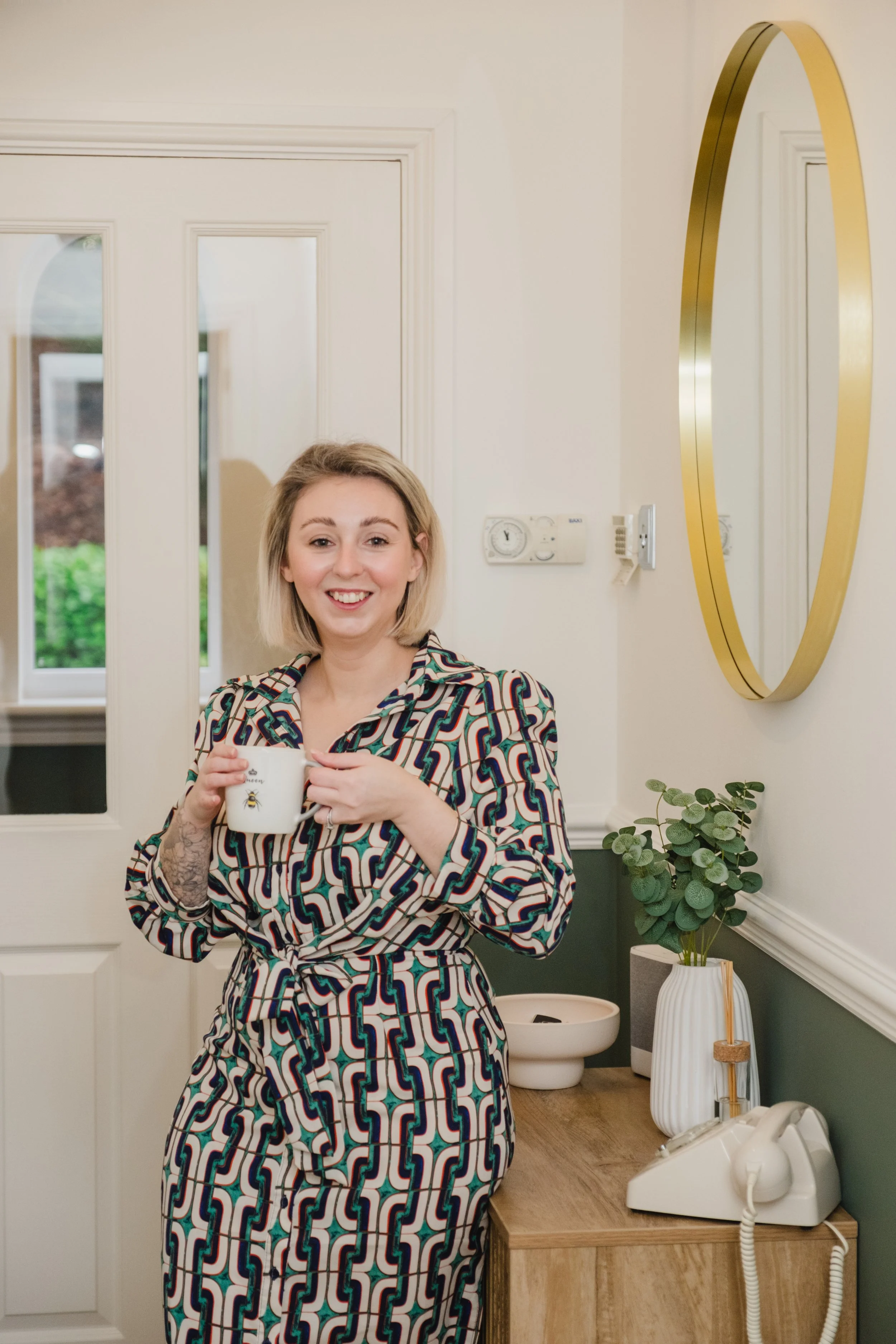 Cassie Marns, Founder of Marns Marketing, wearing a colourful geometric patterned dress, smiling and holding a white mug with a small bee illustration in a room with a wooden side table, a potted plant, a mirror and a vintage telephone.