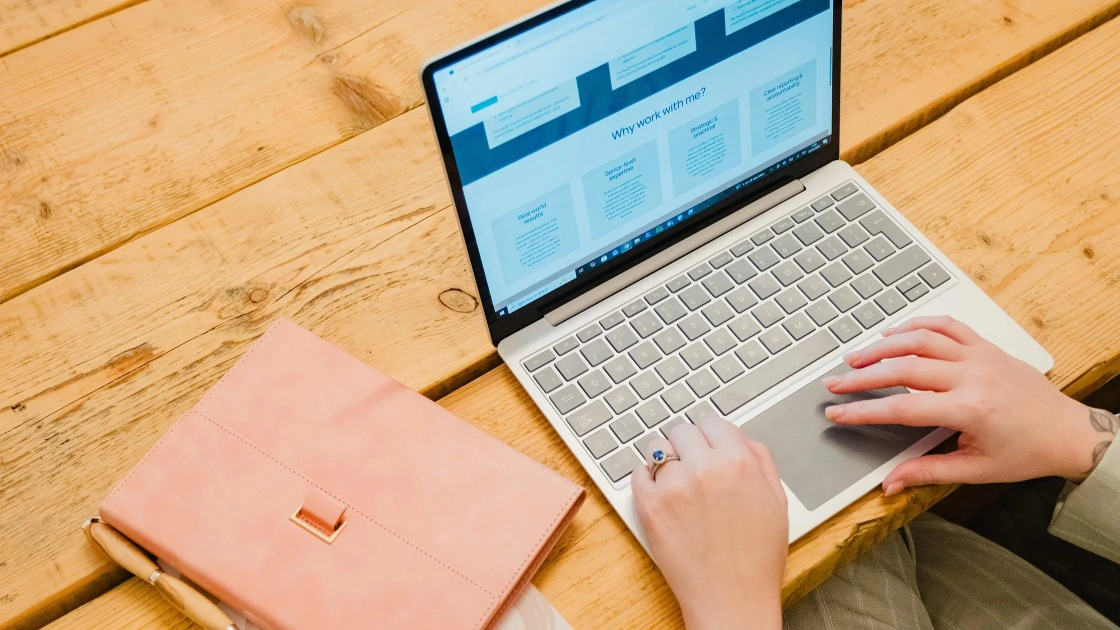 Cassie Marns using a silver laptop on a wooden table with a pink clutch nearby.