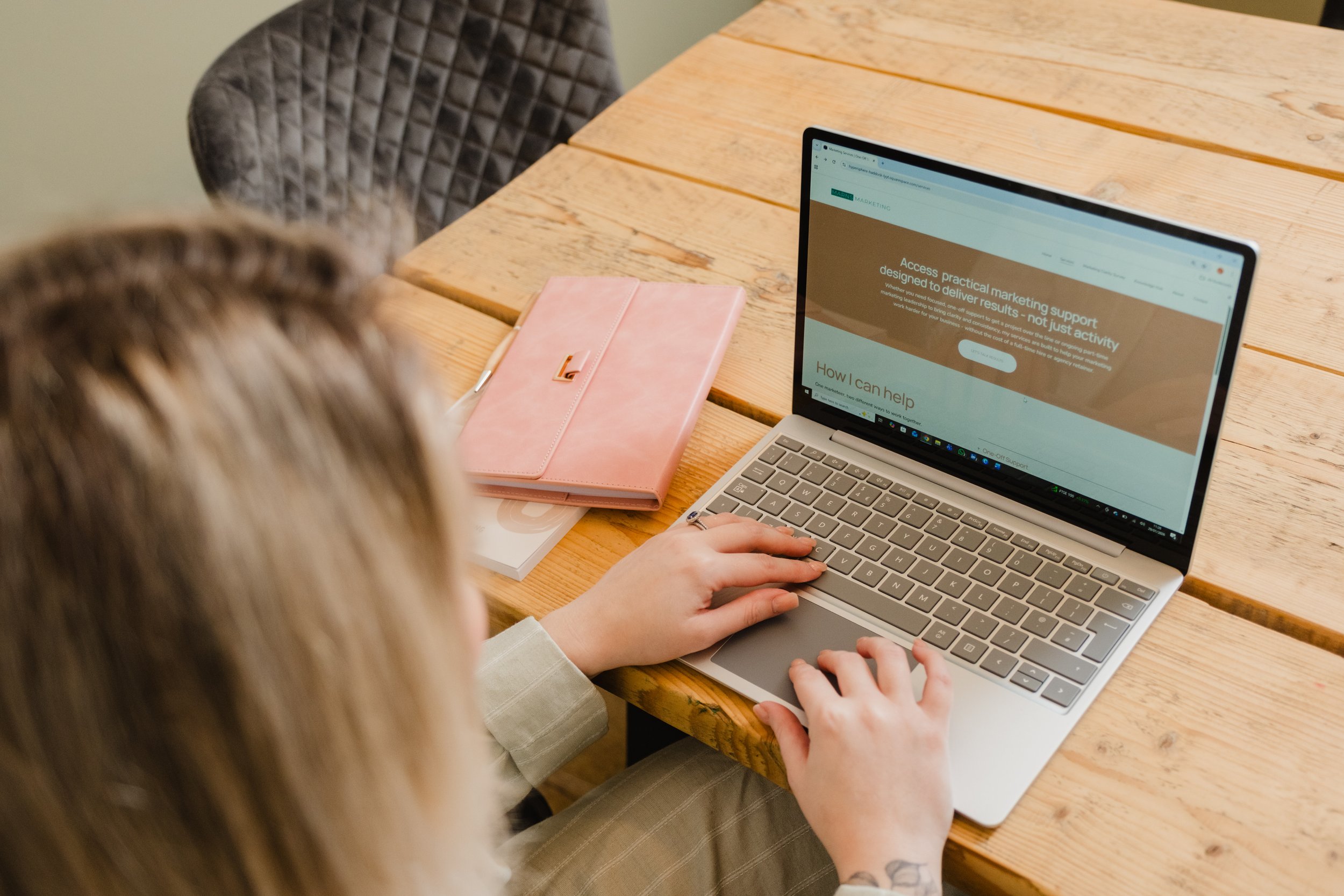 Cassie Marns, Founder of Marns Marketing, using a laptop on a wooden table with a pink notebook and a white stack of papers nearby.