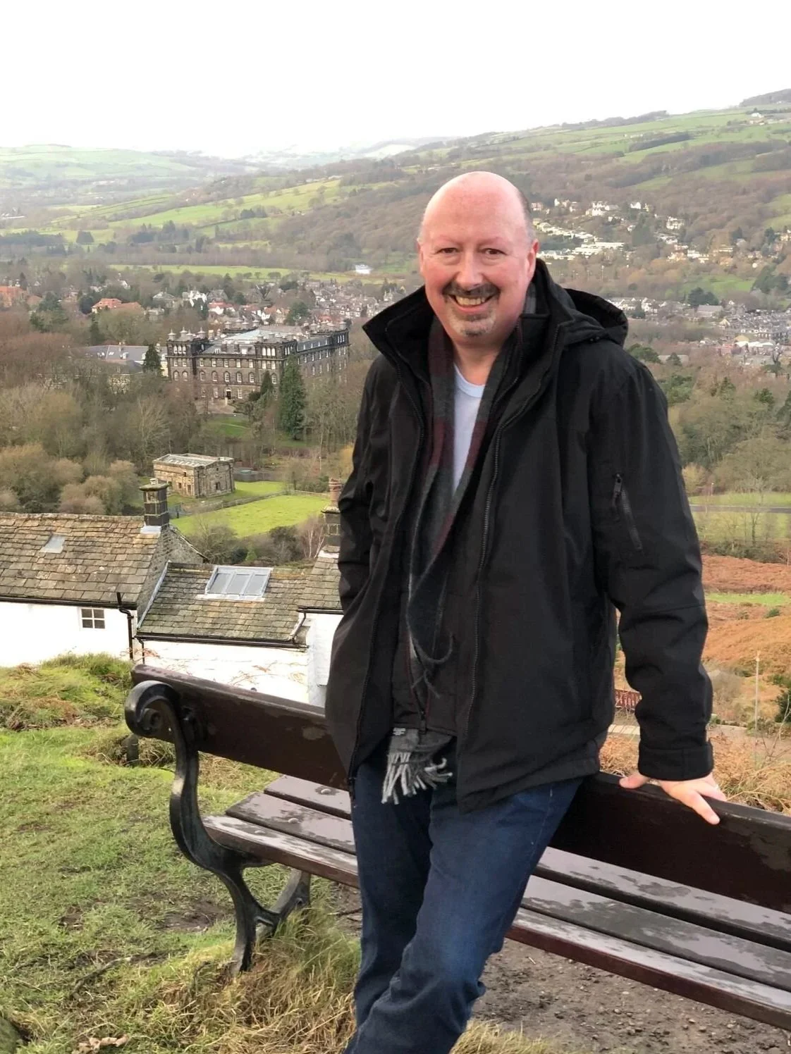 A smiling man standing outdoors next to a park bench with a scenic view of hills, trees, and buildings in the background.