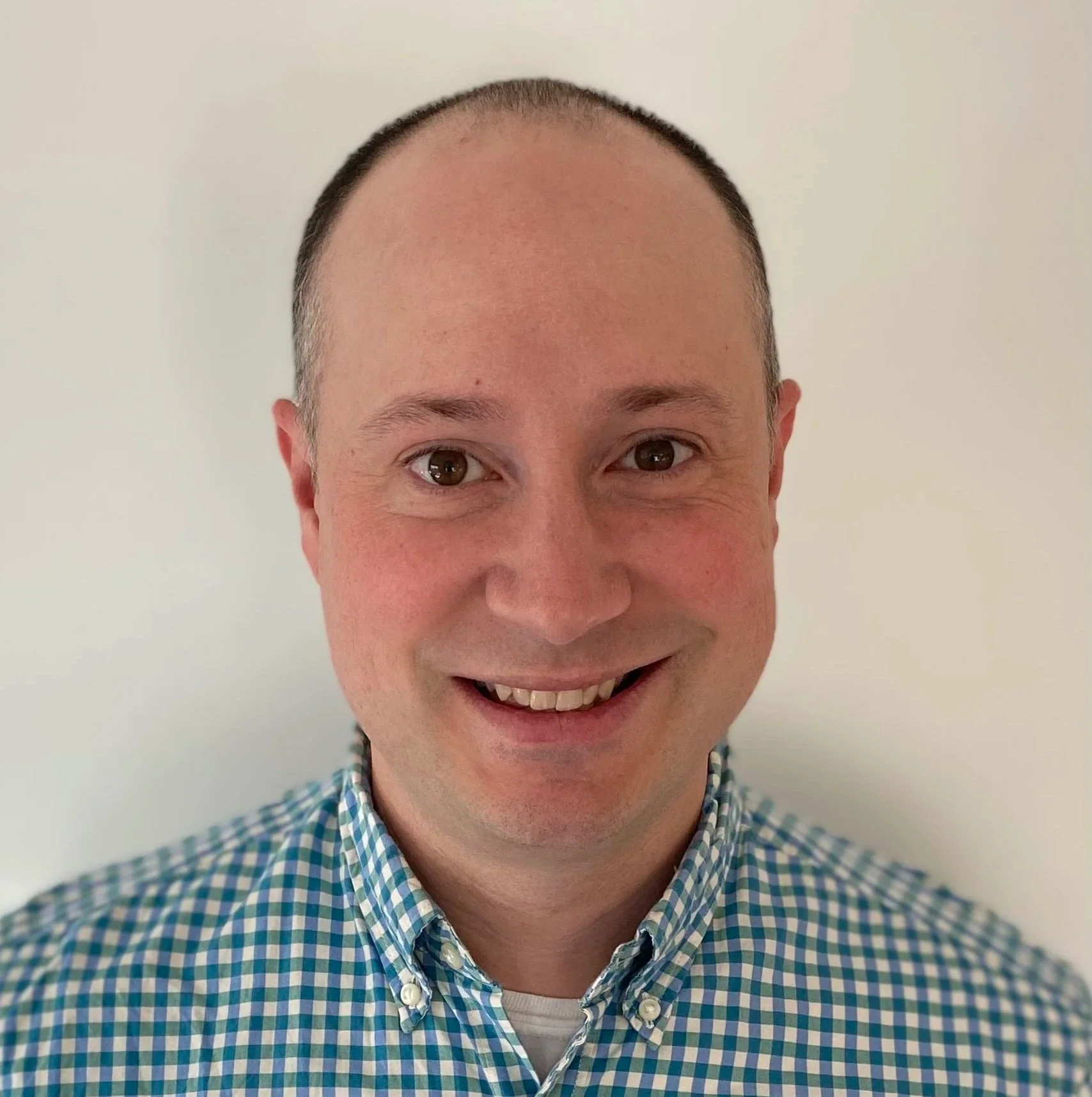 A smiling man with short hair wearing a blue plaid shirt.