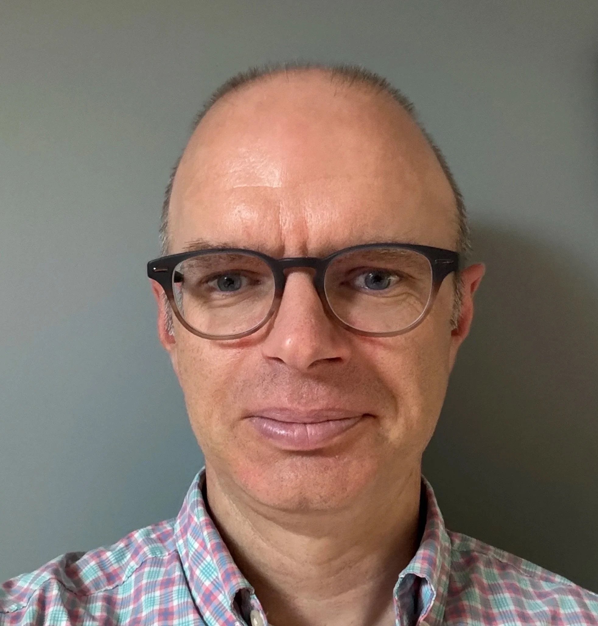 Close-up of a man's face with glasses, wearing a plaid shirt, standing against a plain gray background.