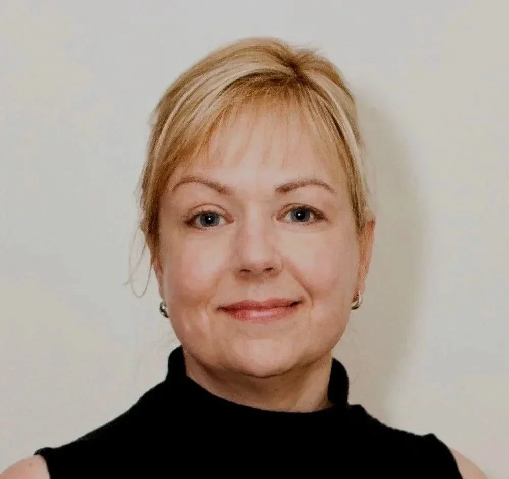 A woman with short blonde hair smiling while looking at the camera, wearing a black top and earrings, against a plain background.
