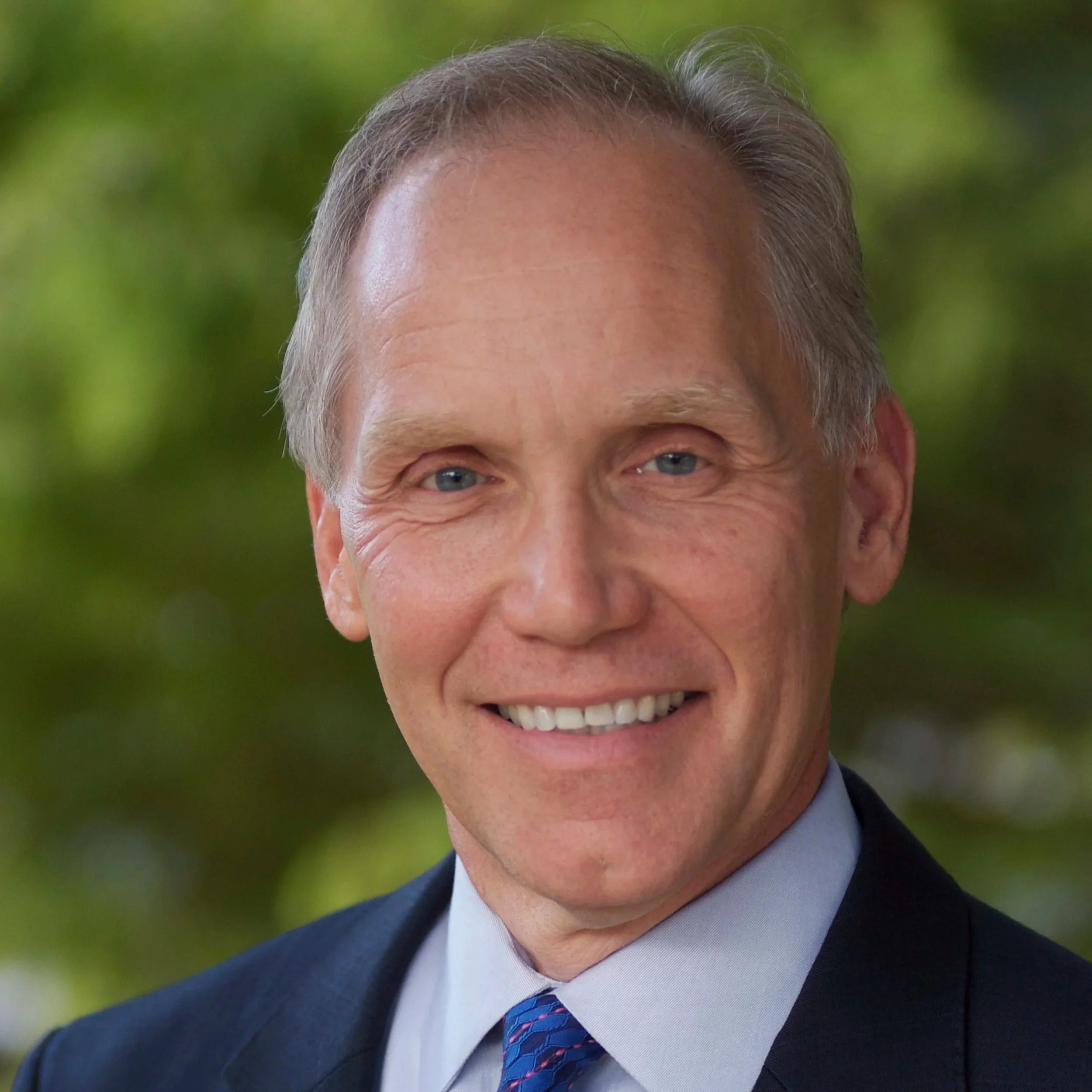 Close-up portrait of a smiling middle-aged man in a suit with a tie, outdoor background with blurred green foliage.