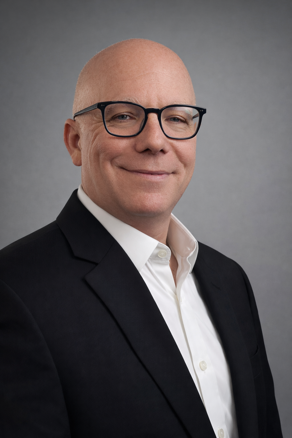 A professional headshot of a middle-aged man with glasses, wearing a black suit and white shirt, smiling slightly, in front of a gray background.
