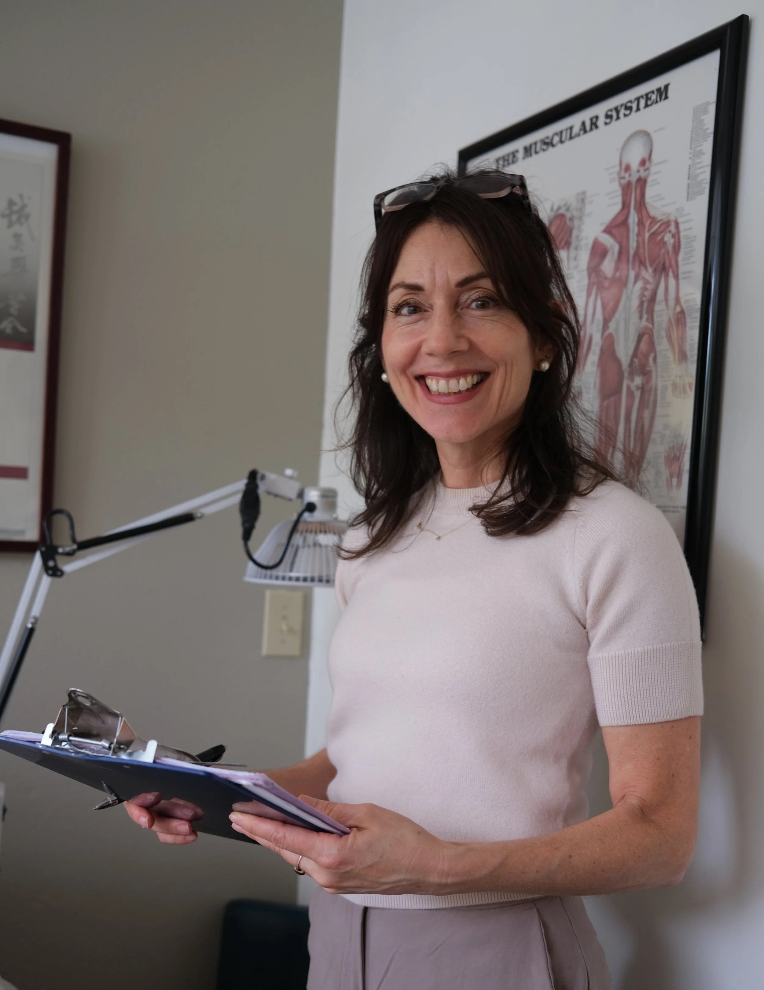 A woman standing and smiling while holding a clipboard with papers, wearing a light-colored short-sleeve top and glasses resting on her head, with drywall and a framed poster of the muscular system in the background.