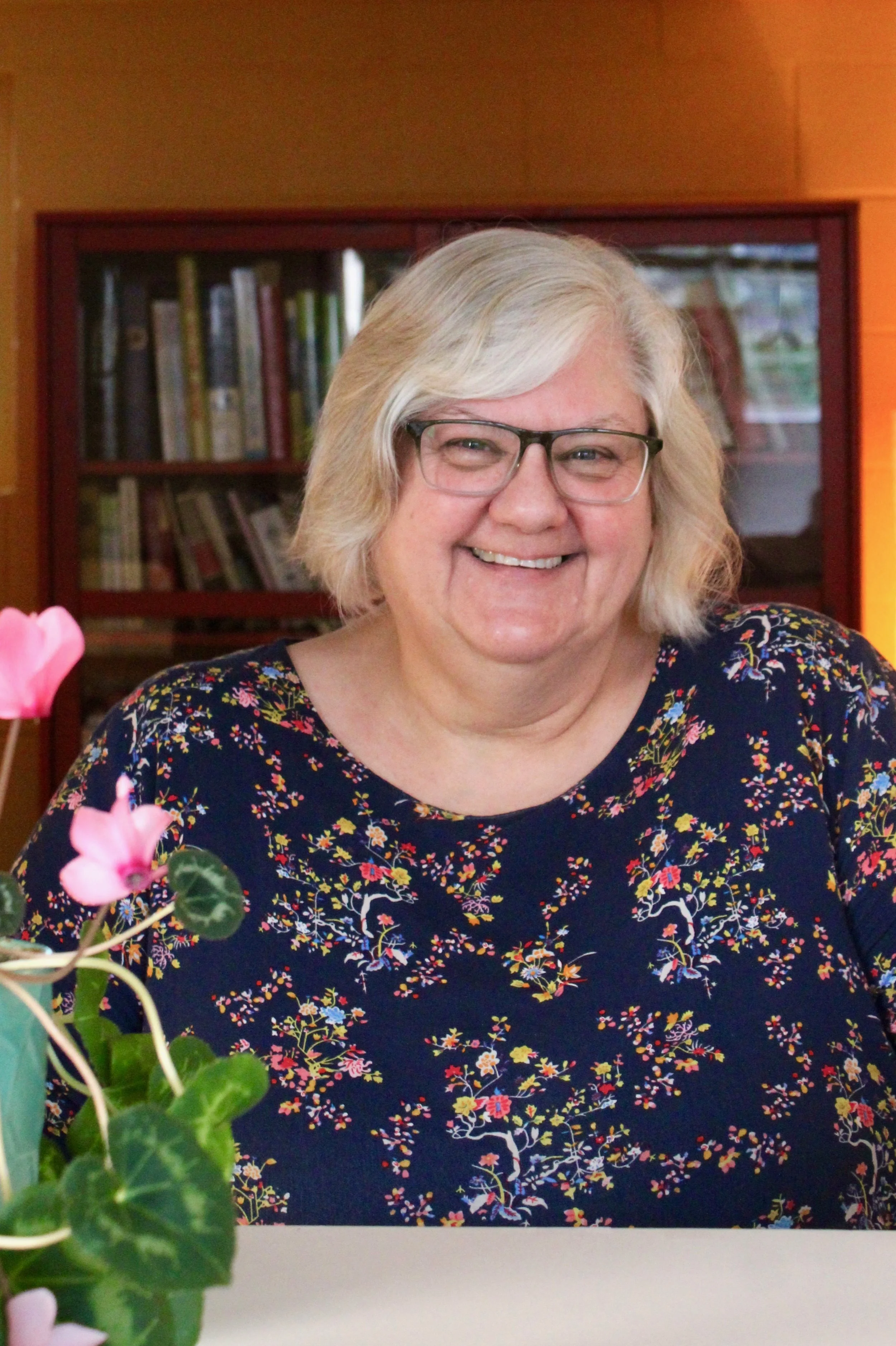 A smiling woman with glasses and short, wavy, gray hair sitting at a table with pink flowers in the foreground. She is wearing a dark floral-patterned top. Behind her is a wooden bookshelf filled with books.