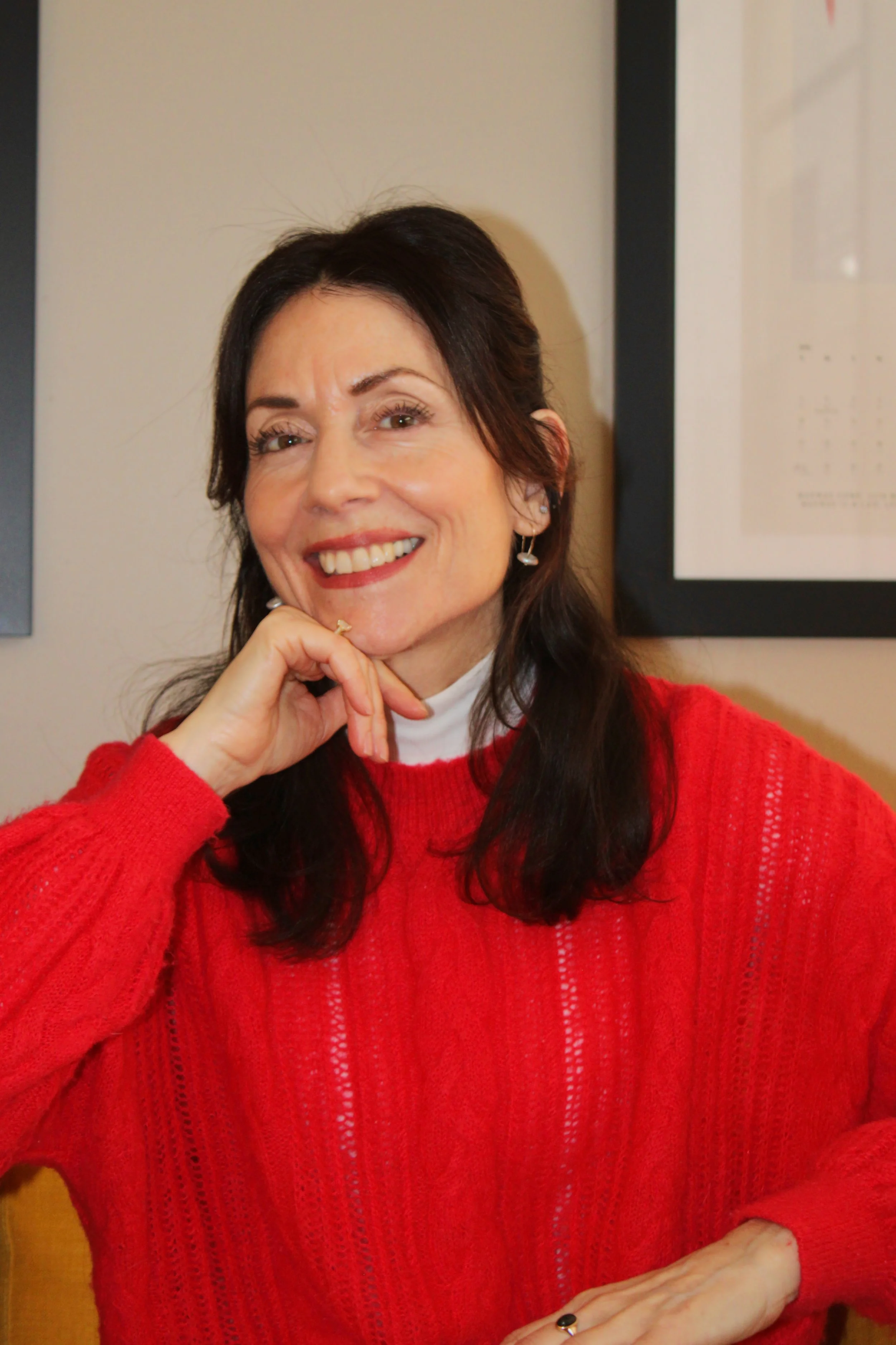 A woman smiling, wearing a red sweater, with dark hair, sitting indoors near framed pictures on the wall.