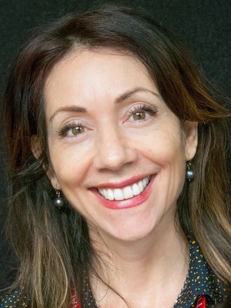 Close-up of smiling woman with brown hair, wearing earrings and a dark patterned shirt, against a black background.