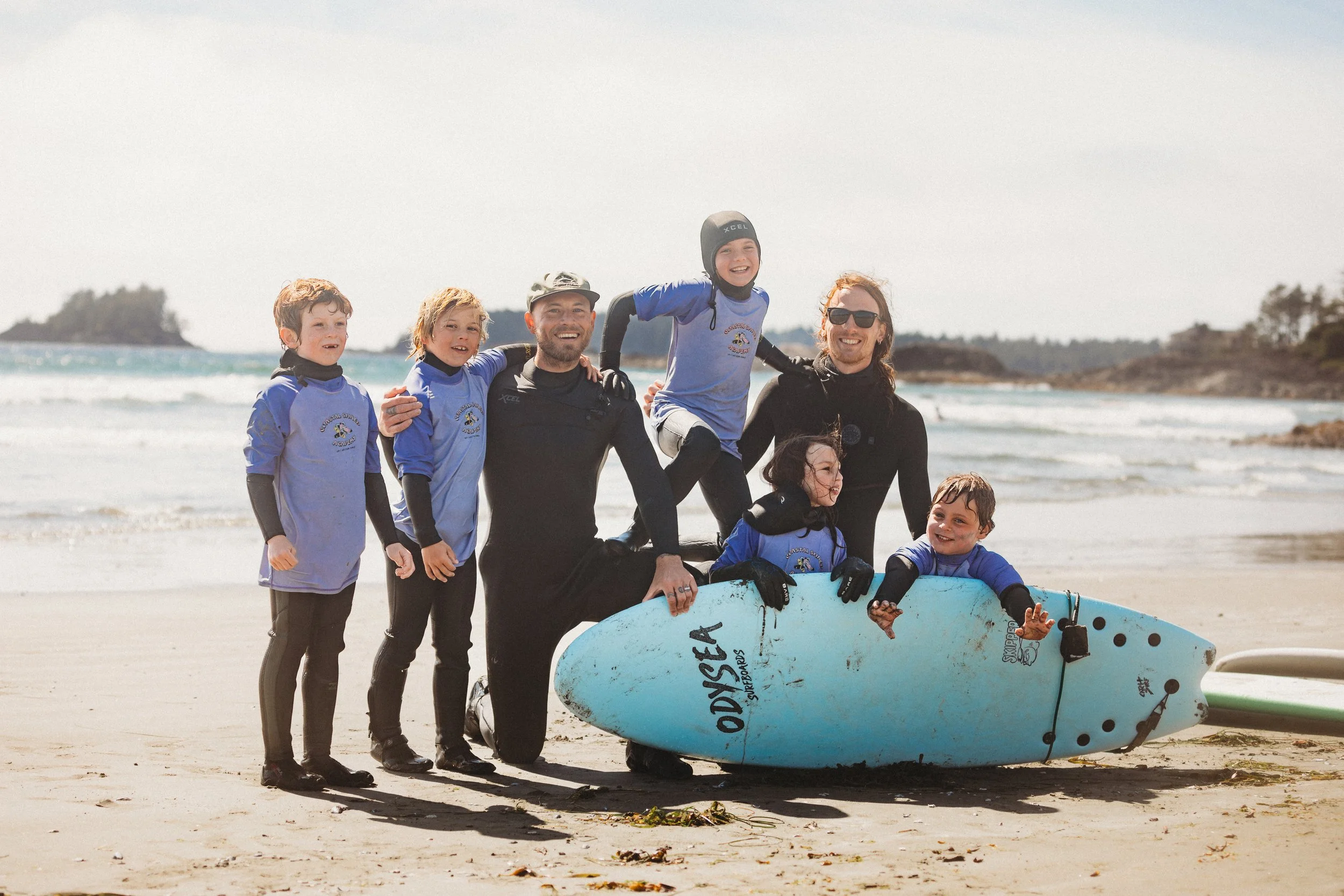 coastal shred academy is known for kids, family and beginner surf lessons. this is instructor aaron and sam with kids having fun surfing and at north chesterman beach tofino, getting a group photo at the end of the surf lesson