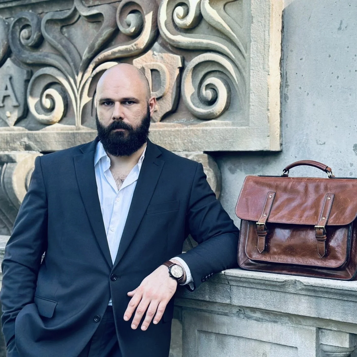Criminal defence lawyer. A man with a beard in a black suit and white shirt, leaning against a stone ledge with a brown leather briefcase on top, standing in front of an ornate stone wall.