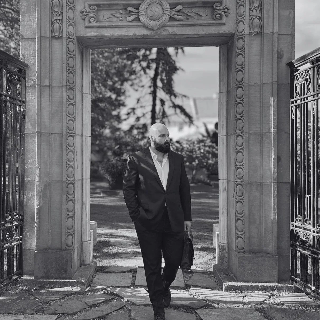 Criminal defence lawyer. Black and white photo of a man with a beard in a suit walking through an ornate stone gate, carrying a bag, with trees and a garden in the background.