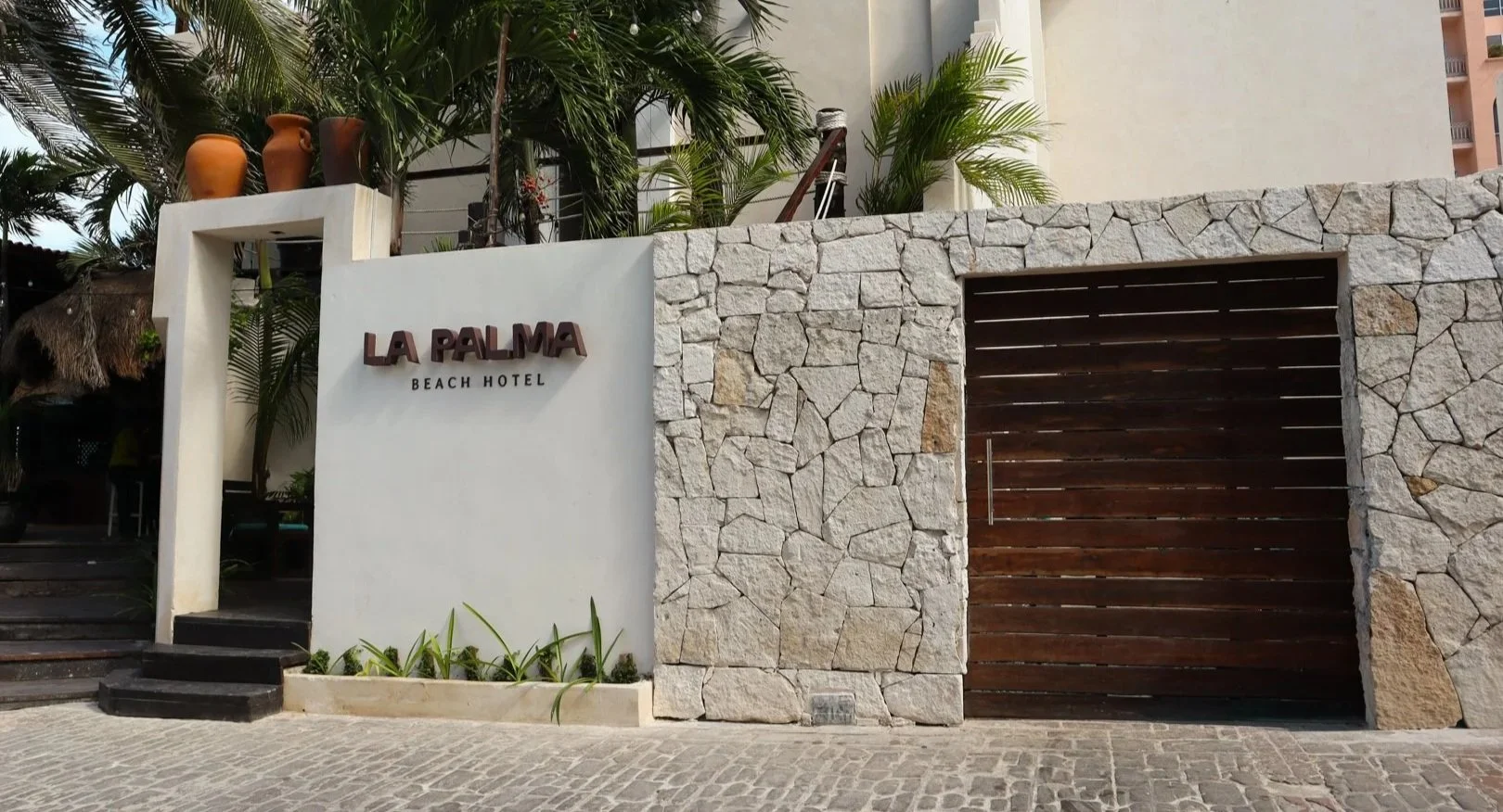 Entrance of La Palma Beach Hotel with stone and wood wall, sign, potted plants, and surrounding foliage.