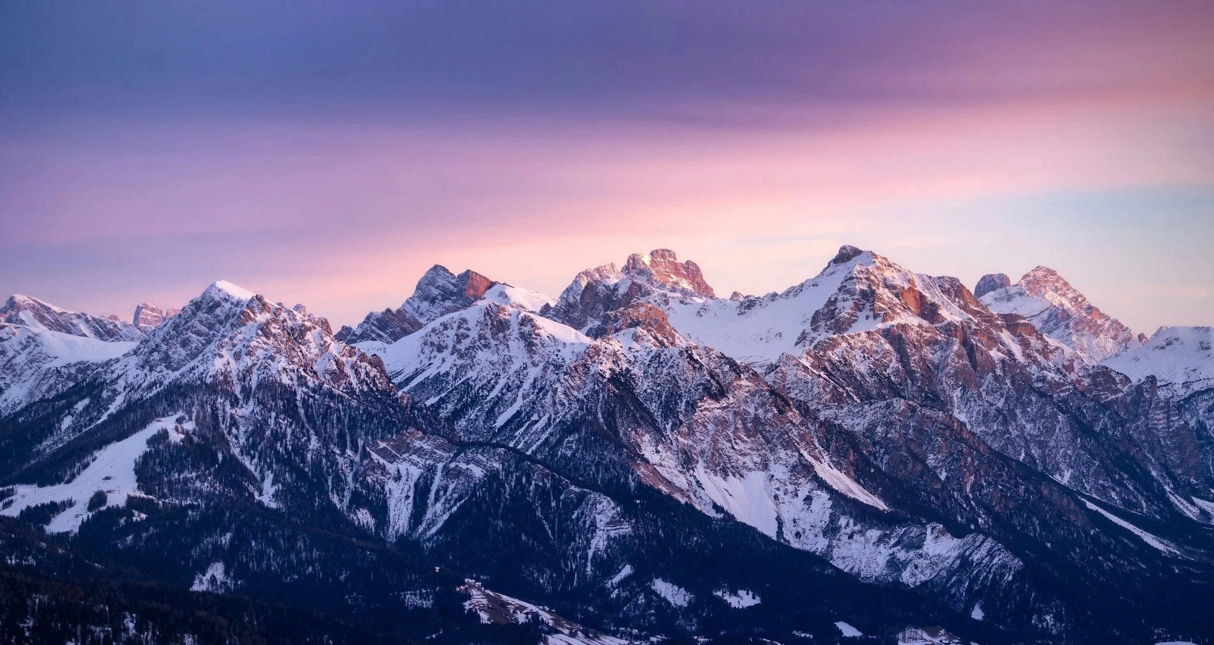 Snow-covered mountain range under pink and purple sky during sunset or sunrise.