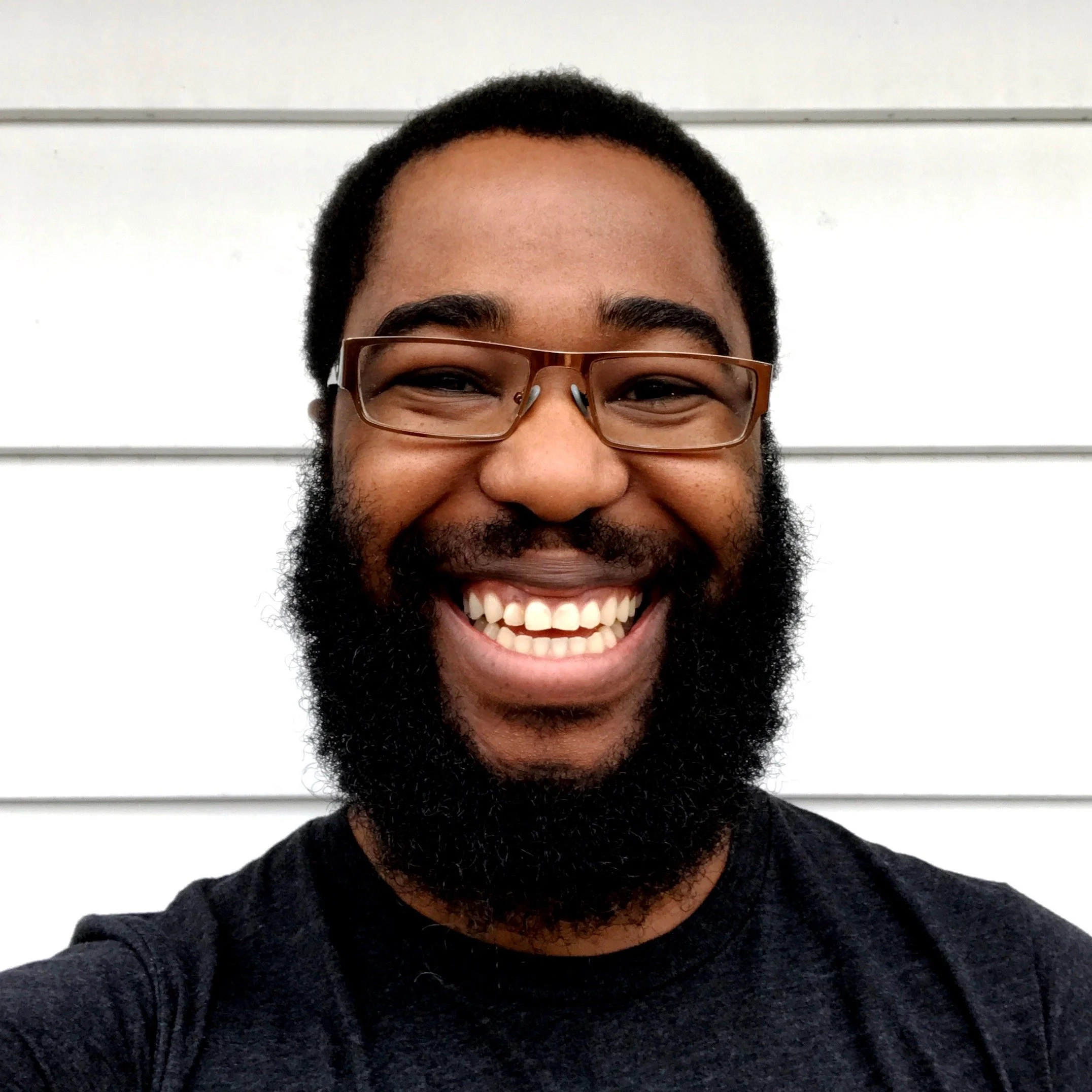 Headshot of Farai Madzima, a Black man wearing glasses and a black t-shirt standing against a white wall.
