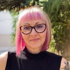 Headshot of Lisa Woodley, a woman with light skin, pink hair, and glasses. She stands outside with leaves in the background.