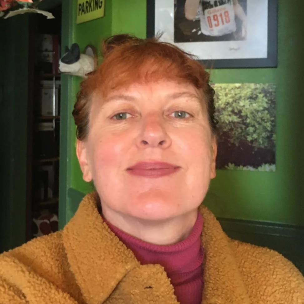 Headshot of Erika Hall, a person with red hair pulled back wearing a camel-colored jacket and red turtleneck. 