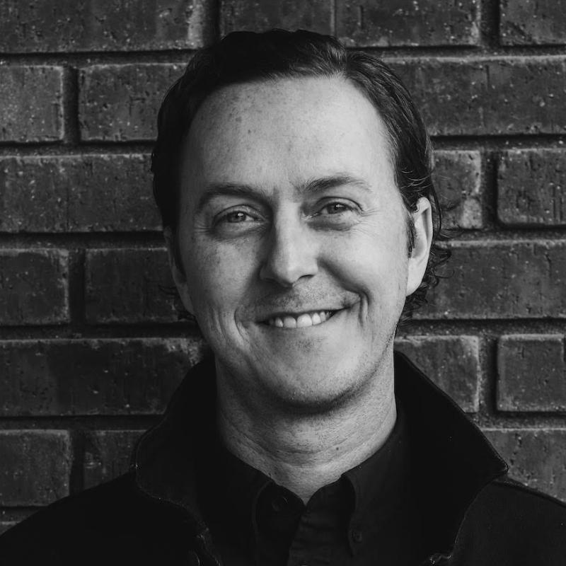 Black and white headshot of Ryan Rumsey, a white man standing against a brick wall.
