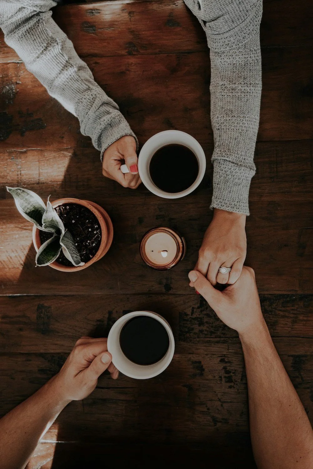 Two people holding coffee cups and holding hands on a wooden table with a candle and a potted plant.