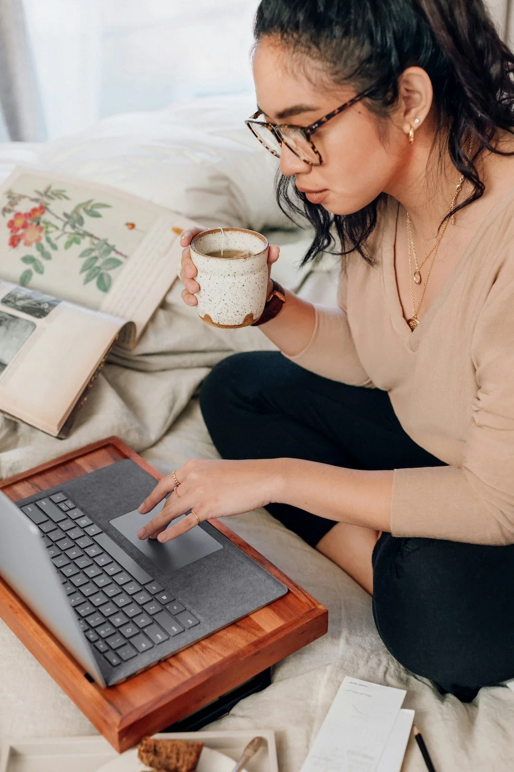 A woman with glasses and jewelry sitting on a bed, working on a laptop placed on a wooden tray, with open books and a cup of tea nearby.