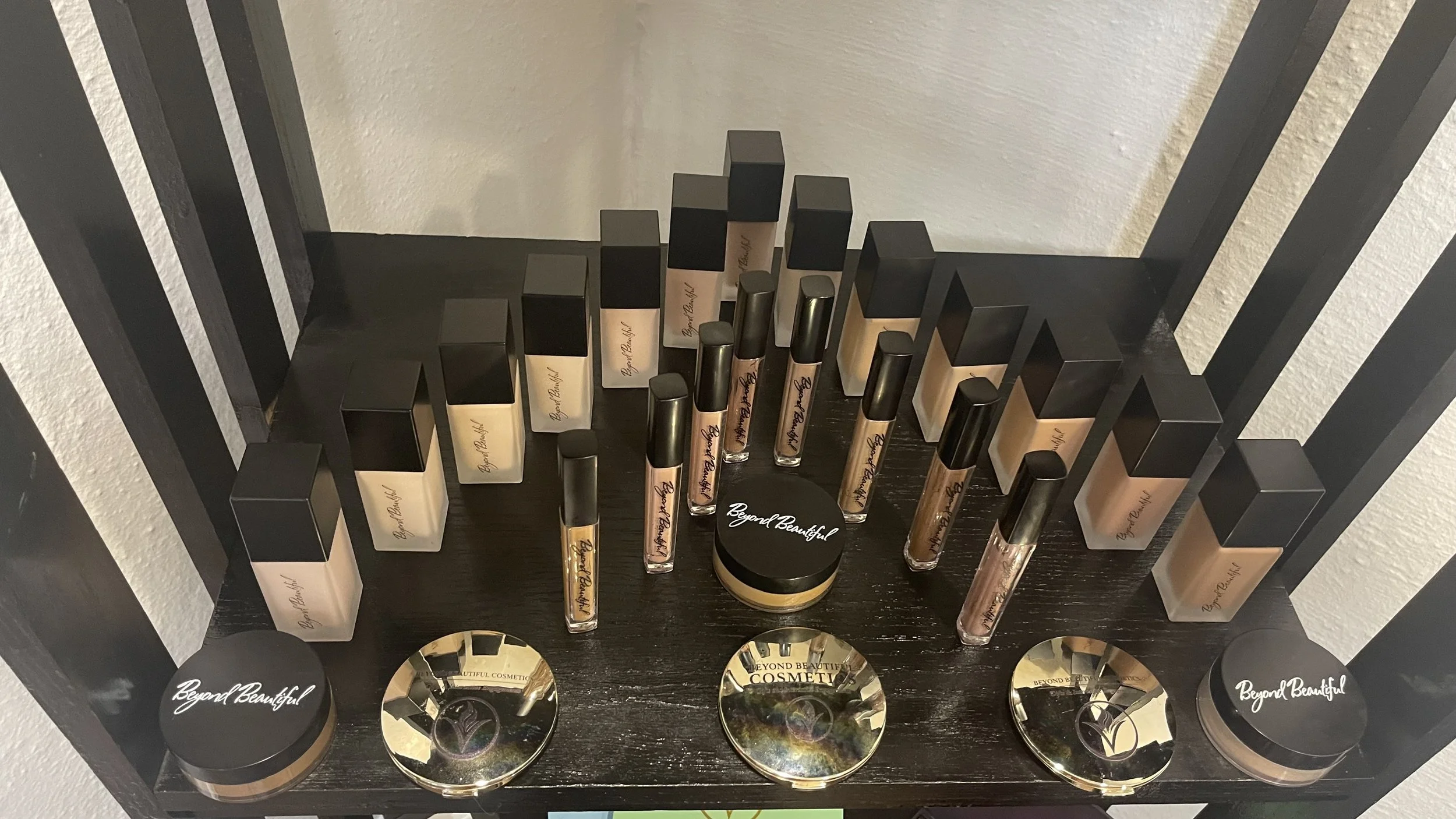 Display of various makeup products including liquid foundation bottles, lip glosses, and powders on a black shelf with gold accents and round mirrors with "Beyond Beautiful" written on them.