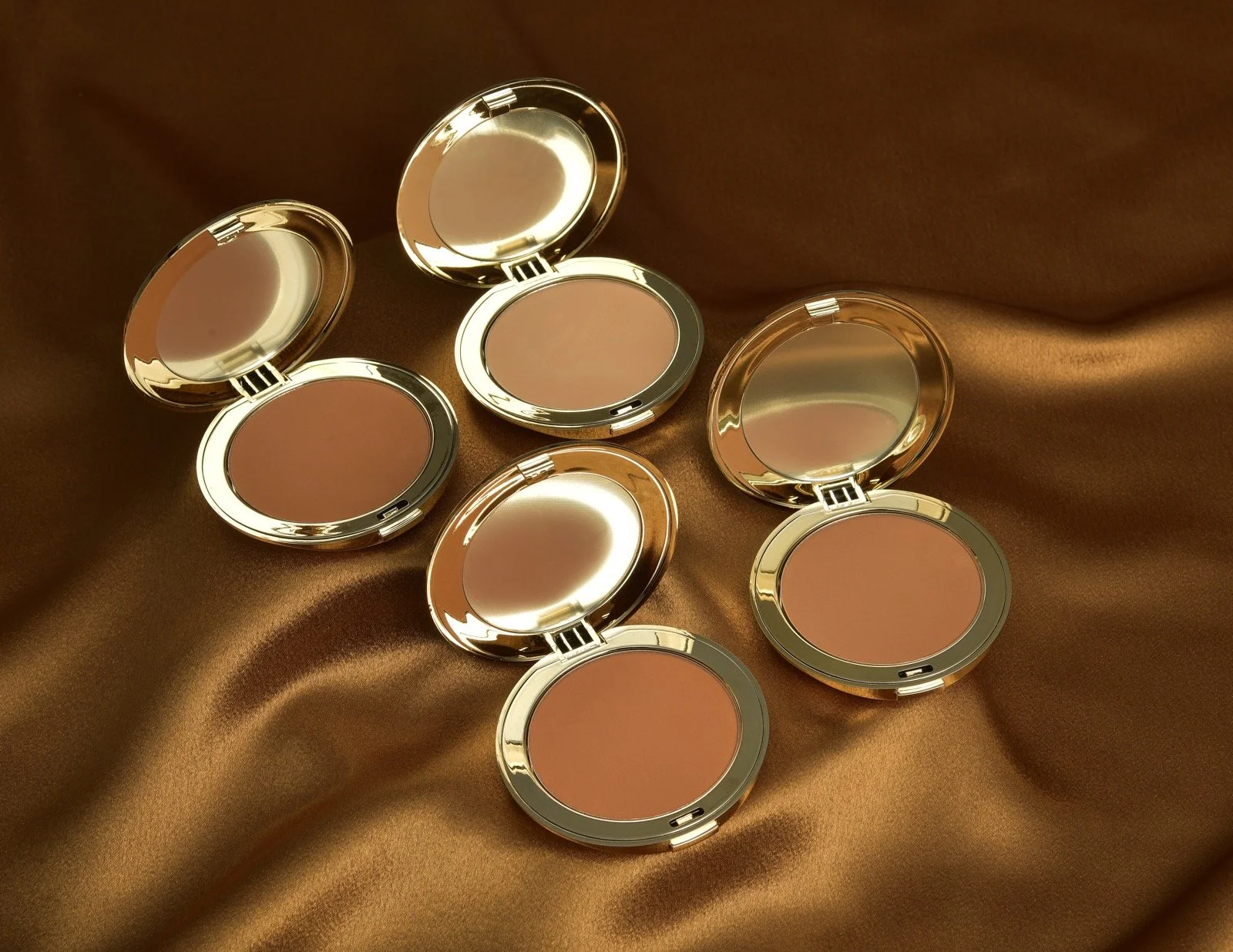 Five compact powder containers with gold-colored cases, each with a reflective mirror inside the lid, placed on a satin brown fabric background.