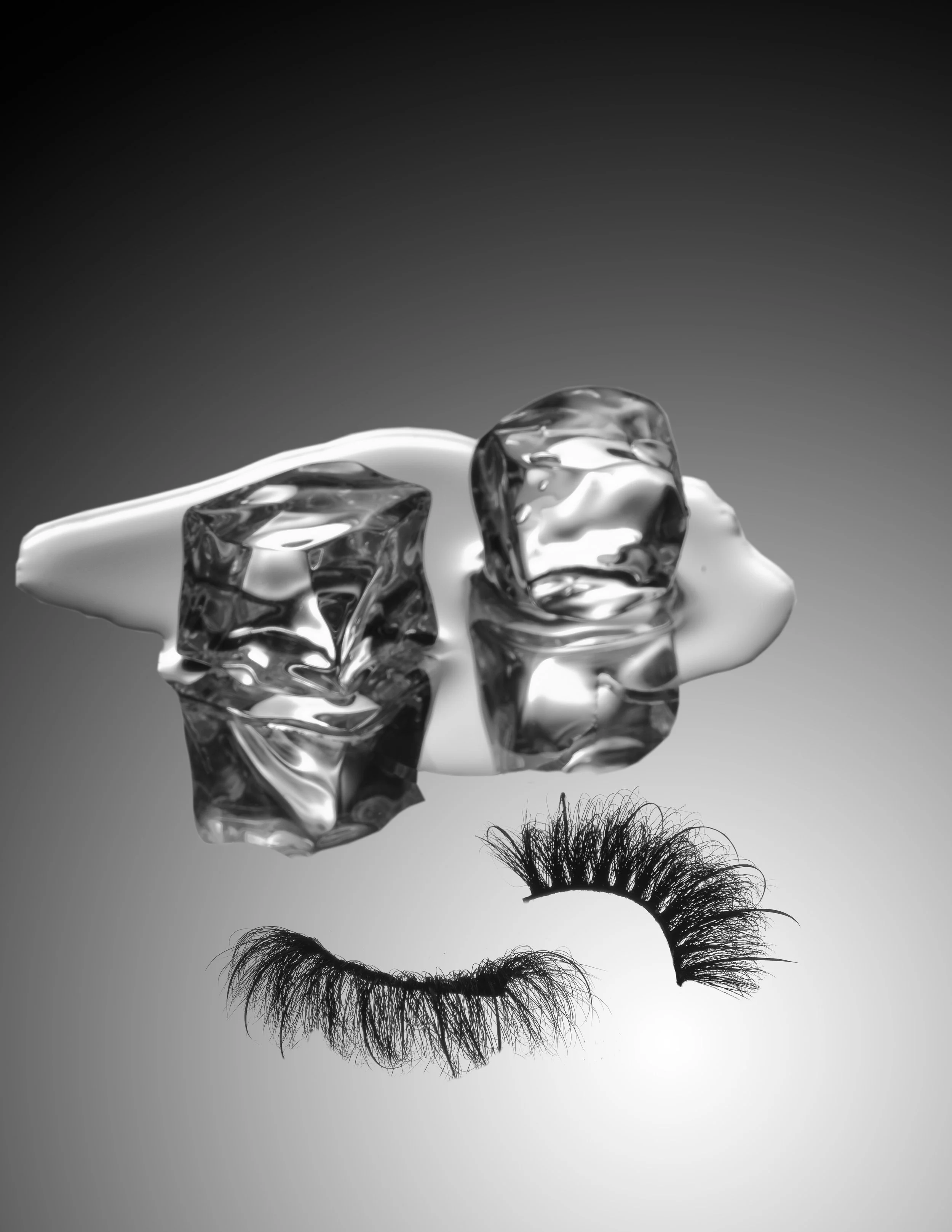 Black and white photo of a person's eyelash makeup, with the upper eyelashes and detached eye lashes on a reflective surface, and ice cubes above.